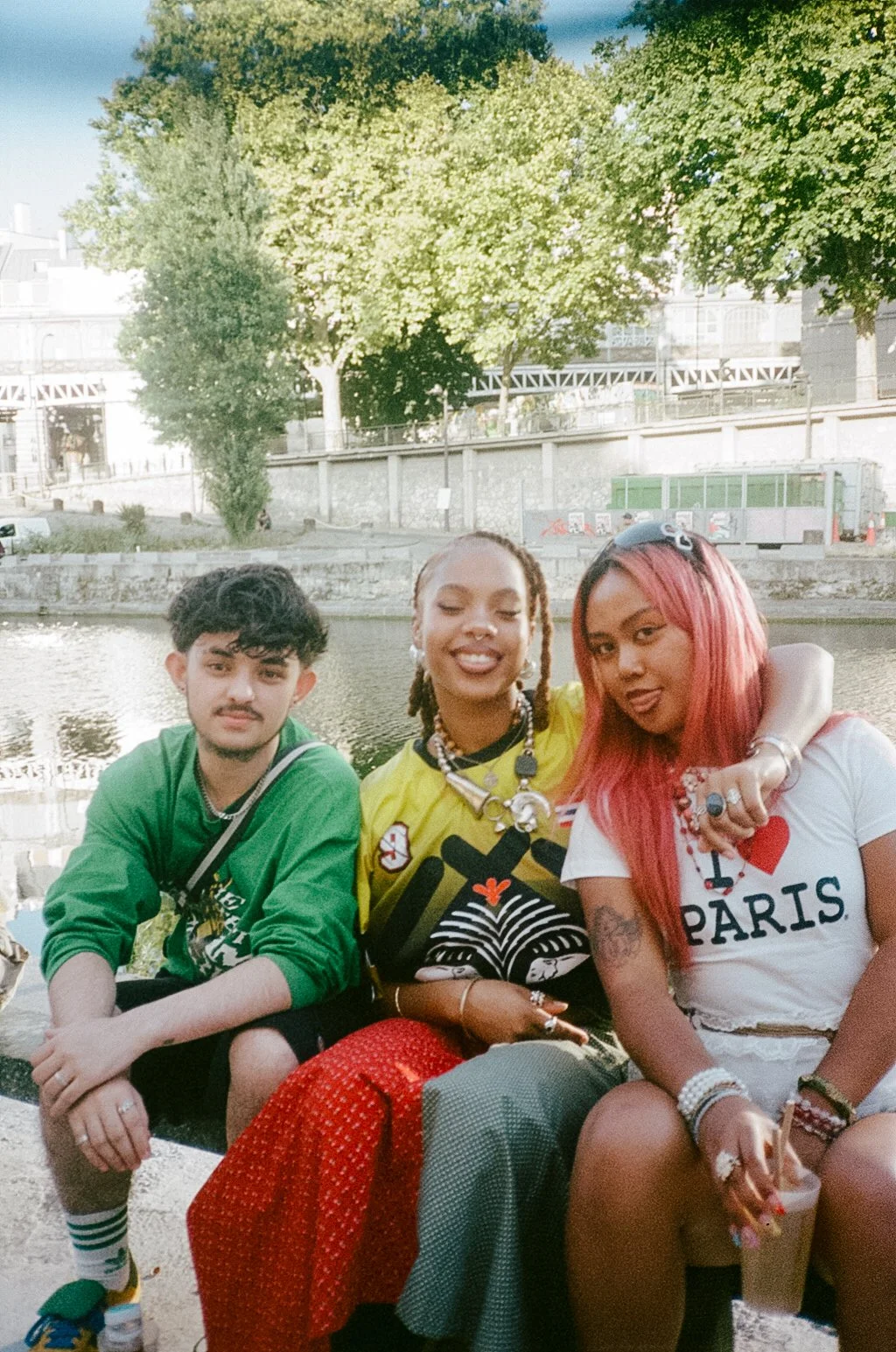Three young people sitting by a river, smiling and enjoying a sunny day, with trees and a building in the background.
