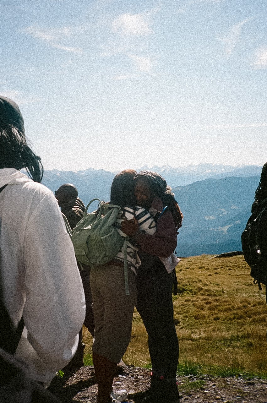 Two women hugging outdoors on a mountain hike, with scenic mountains and a blue sky in the background, surrounded by other hikers.