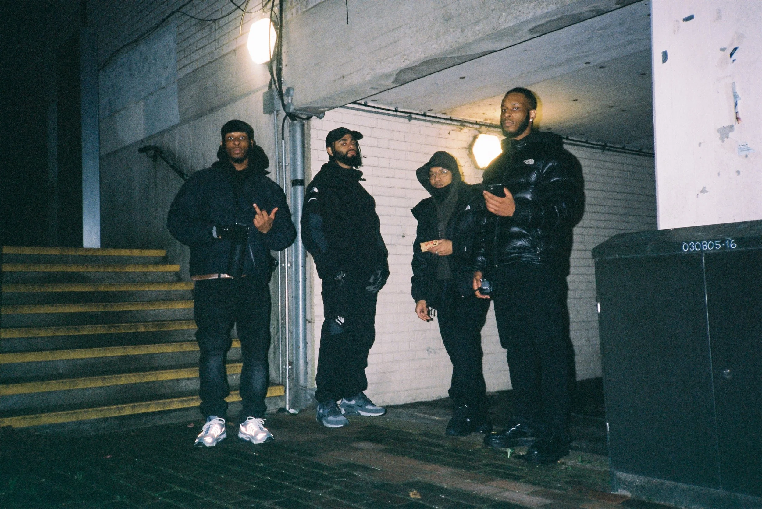 Four men standing outdoors at night near a staircase and a white brick wall, with two lights illuminating the scene. They are dressed in dark clothing, and one man is holding a smartphone.