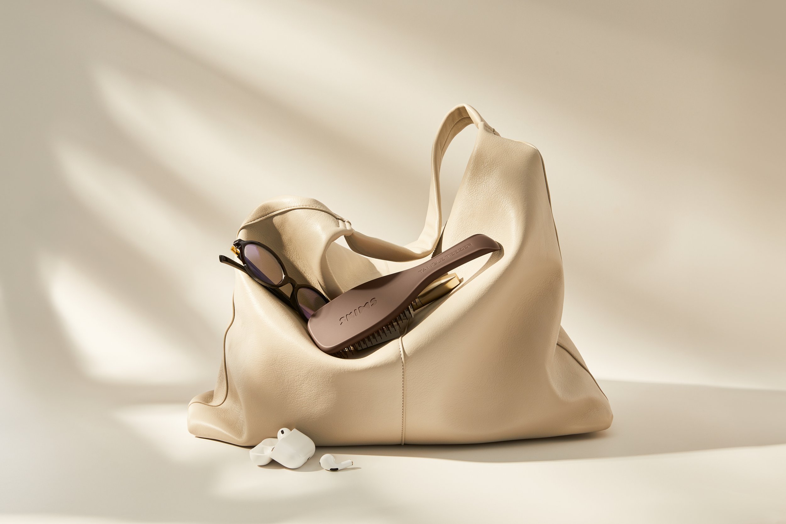 A beige slouch handbag containing reading glasses, a dark brown hairbrush, and white wireless earbuds, all placed on a cream surface with shadow lighting.