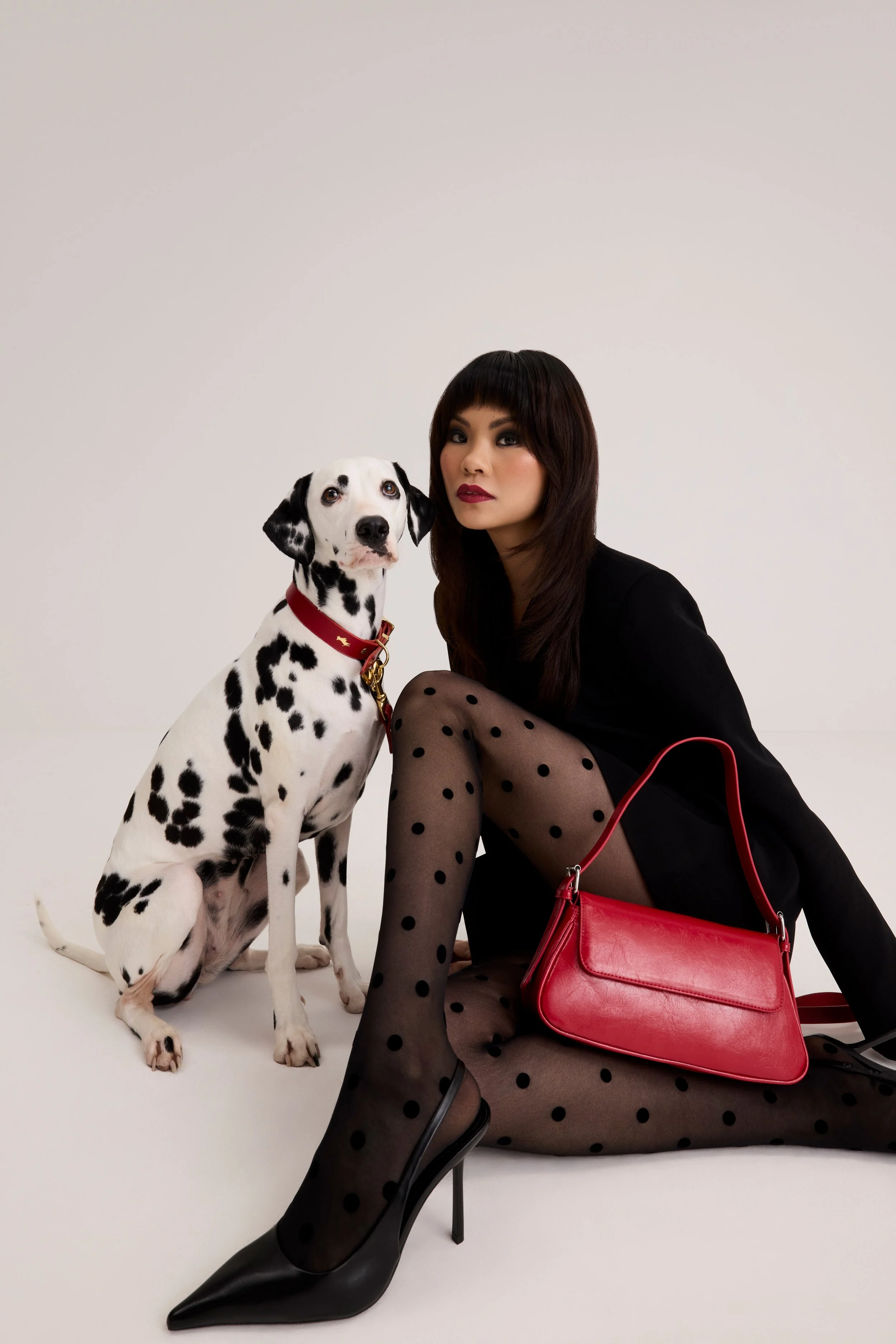 A woman with dark hair and eye makeup sitting on the floor next to a Dalmatian dog. The woman is wearing a black dress, polka dot stockings, and black high heels. A red handbag is on her lap. The background is plain white.