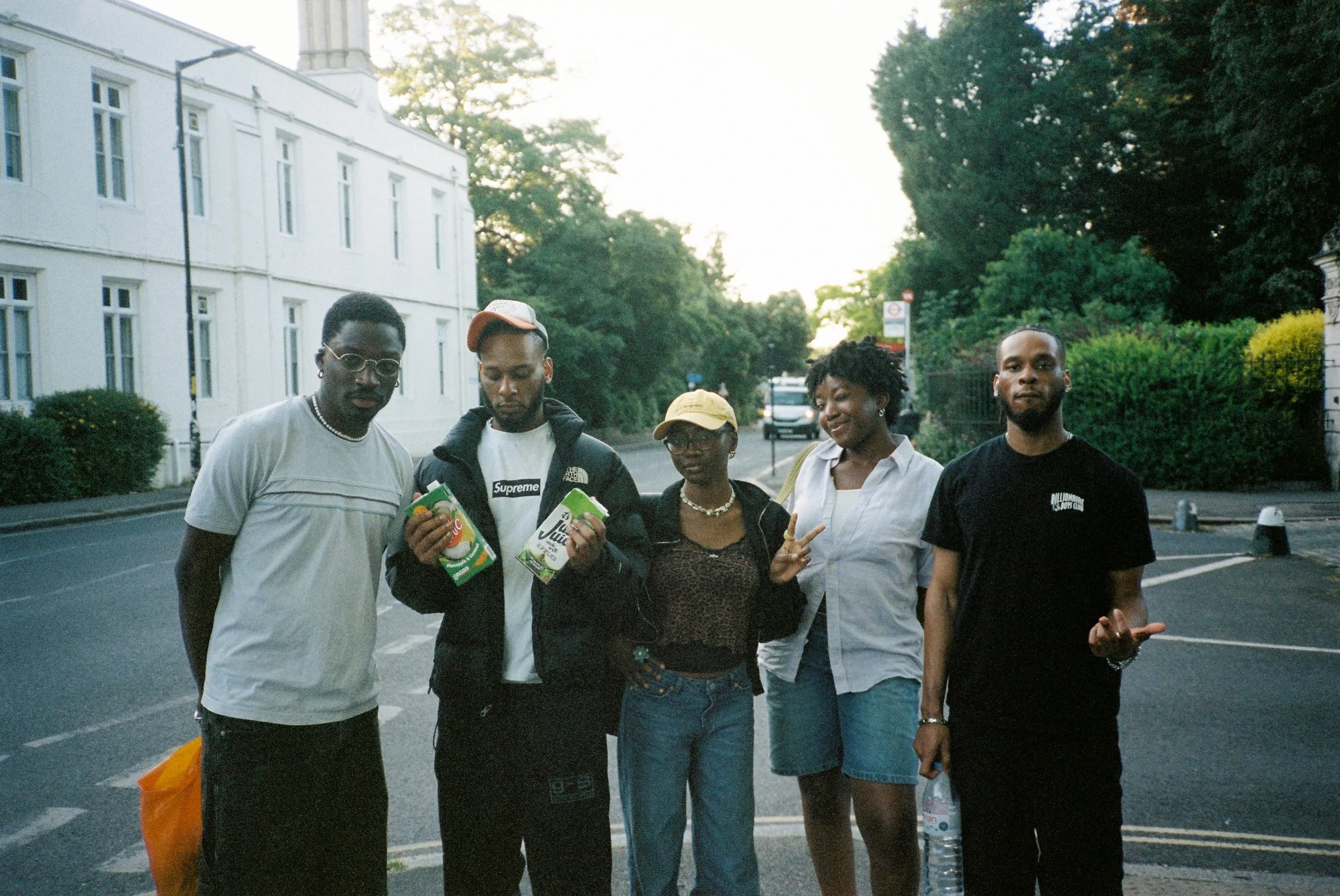 Group of five young adults standing outdoors on a street, holding juice cartons, with trees and white building in the background.