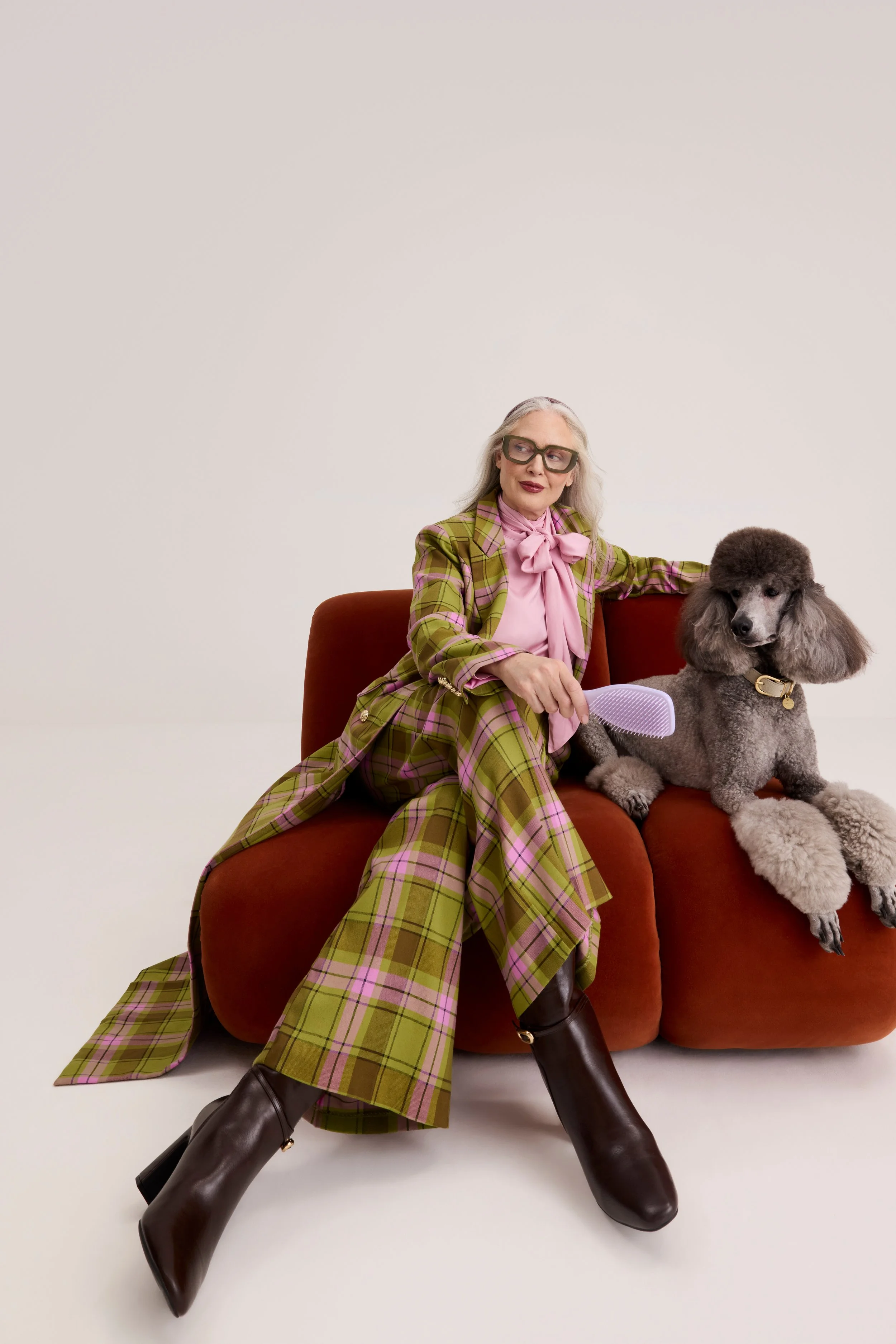 An older woman sitting on a red velvet sofa with a poodle dog beside her. She is wearing a green and pink plaid suit with a pink blouse, black boots, and glasses. The woman is combing the poodle, which has curly gray fur.