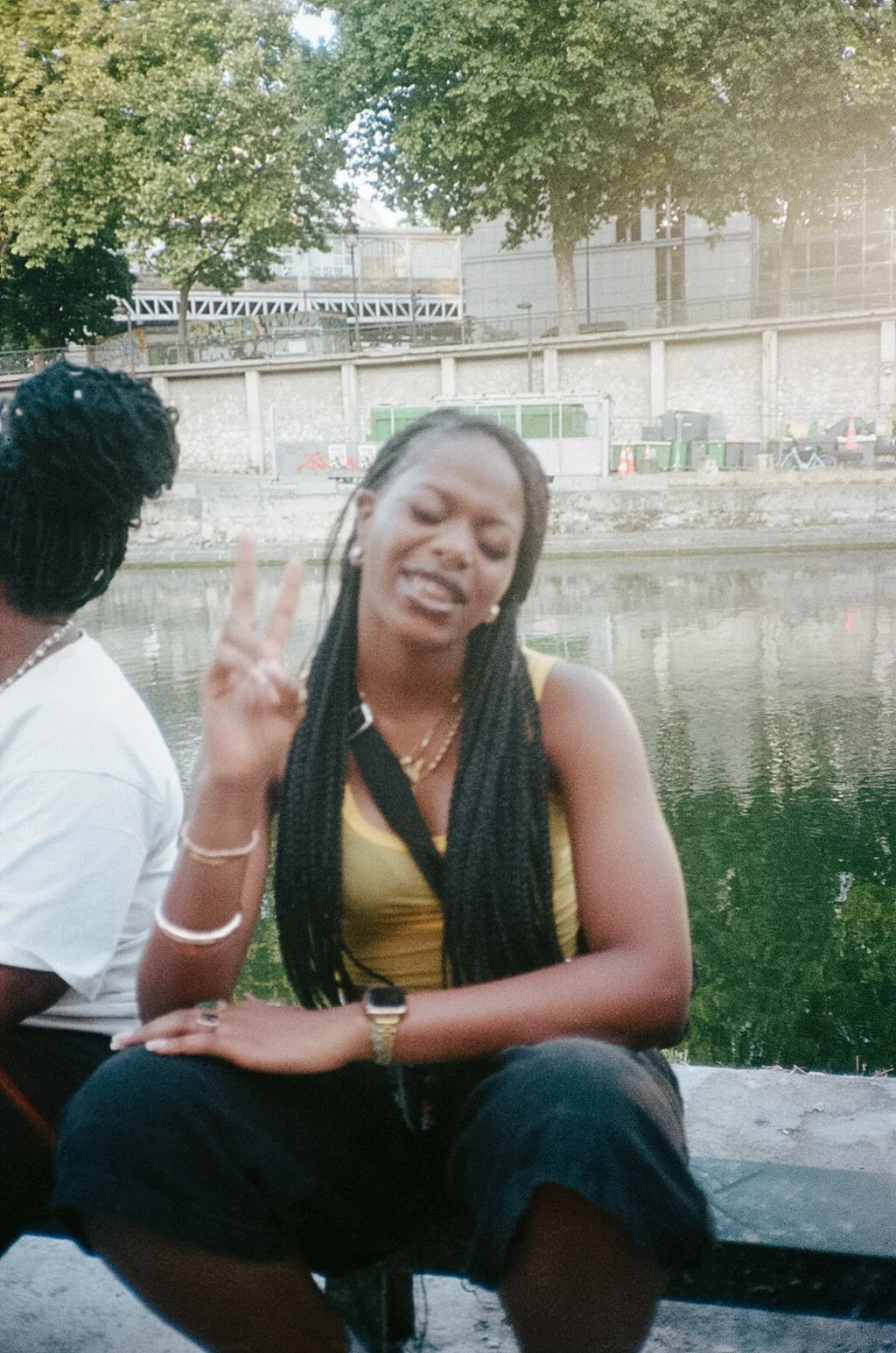 A young woman with long braided hair making a peace sign and smiling at the camera near a river, with trees, a stone wall, and a building in the background.