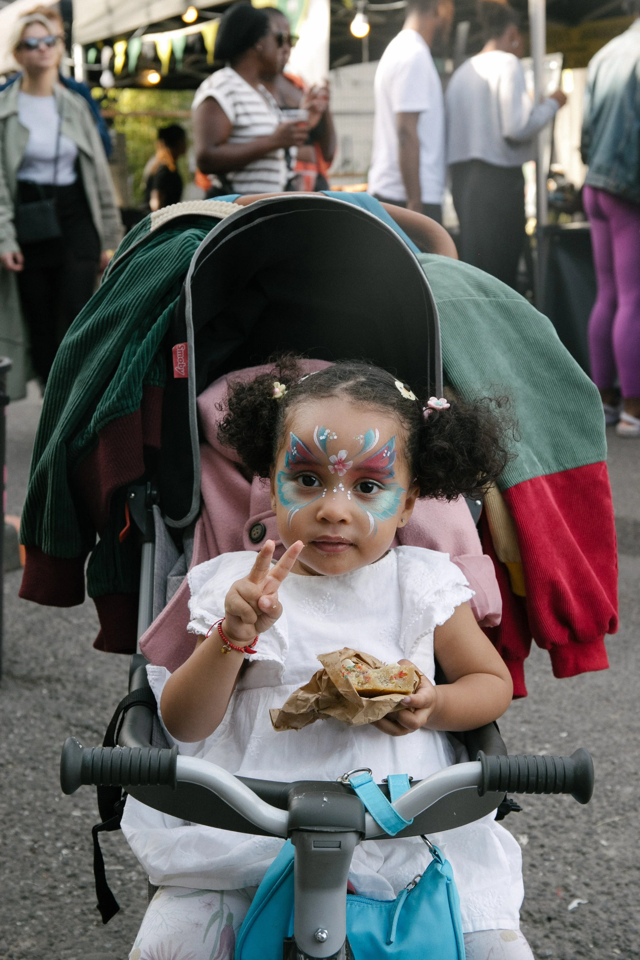 Young girl with face paint of a butterfly, sitting in a buggy, holding a snack wrapped in paper, and making a peace sign gesture with her fingers at an outdoor event.