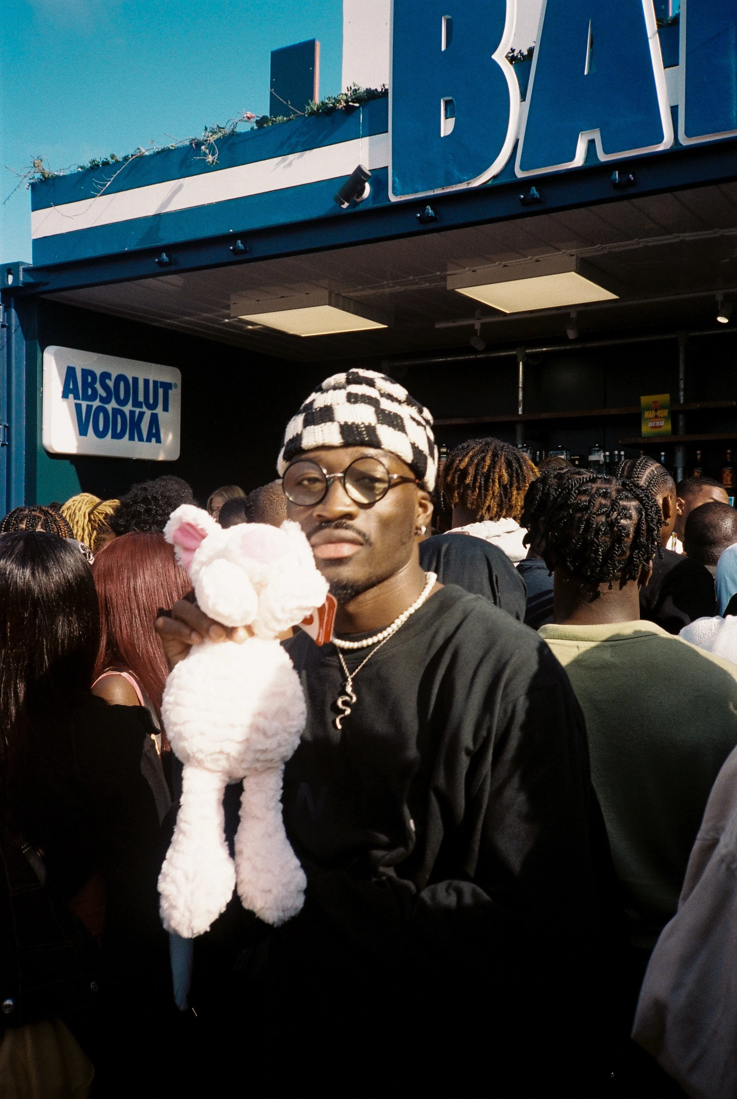 A young man wearing glasses and a black and white checkered beanie hat holds a large white plush bear in a crowded outdoor setting. In the background, there are many people, a sign for Absolut Vodka, and a blue sign with partially visible white lette