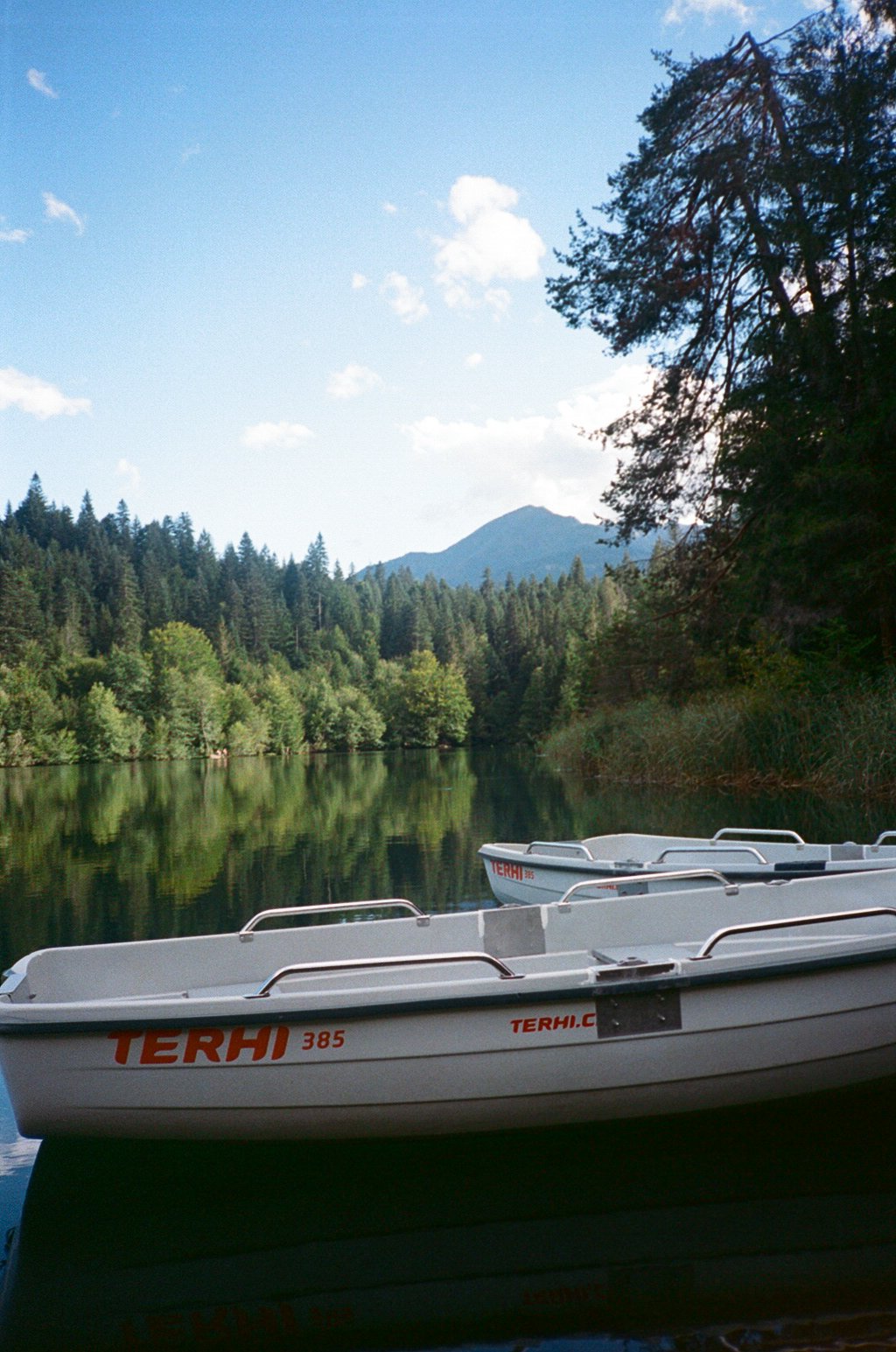 Two boats floating on a calm river surrounded by tall green trees and mountain in the background on a clear day.