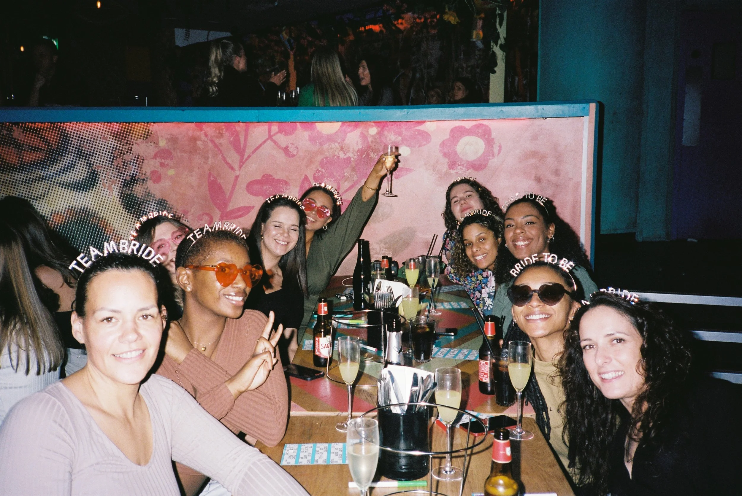 Group of women celebrating at a party, some wearing 'Bride to Be' headbands, with drinks on the table in a dimly lit venue with colorful decor in the background.
