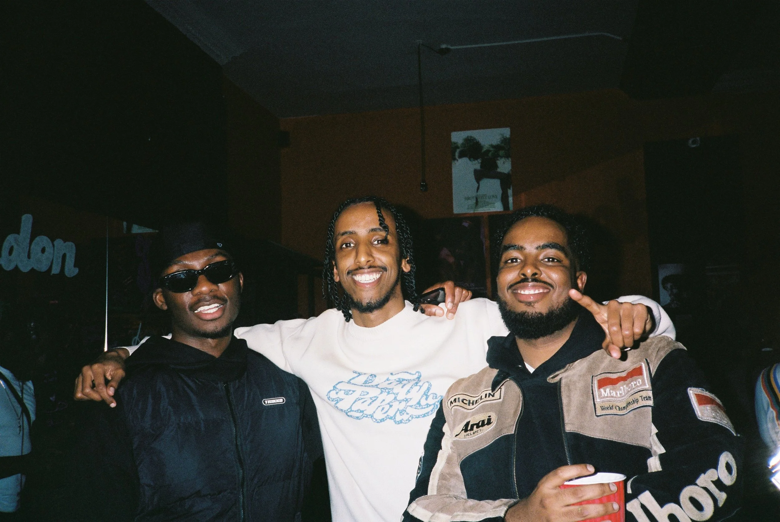 Three people smiling and standing close together in a dimly lit room. 