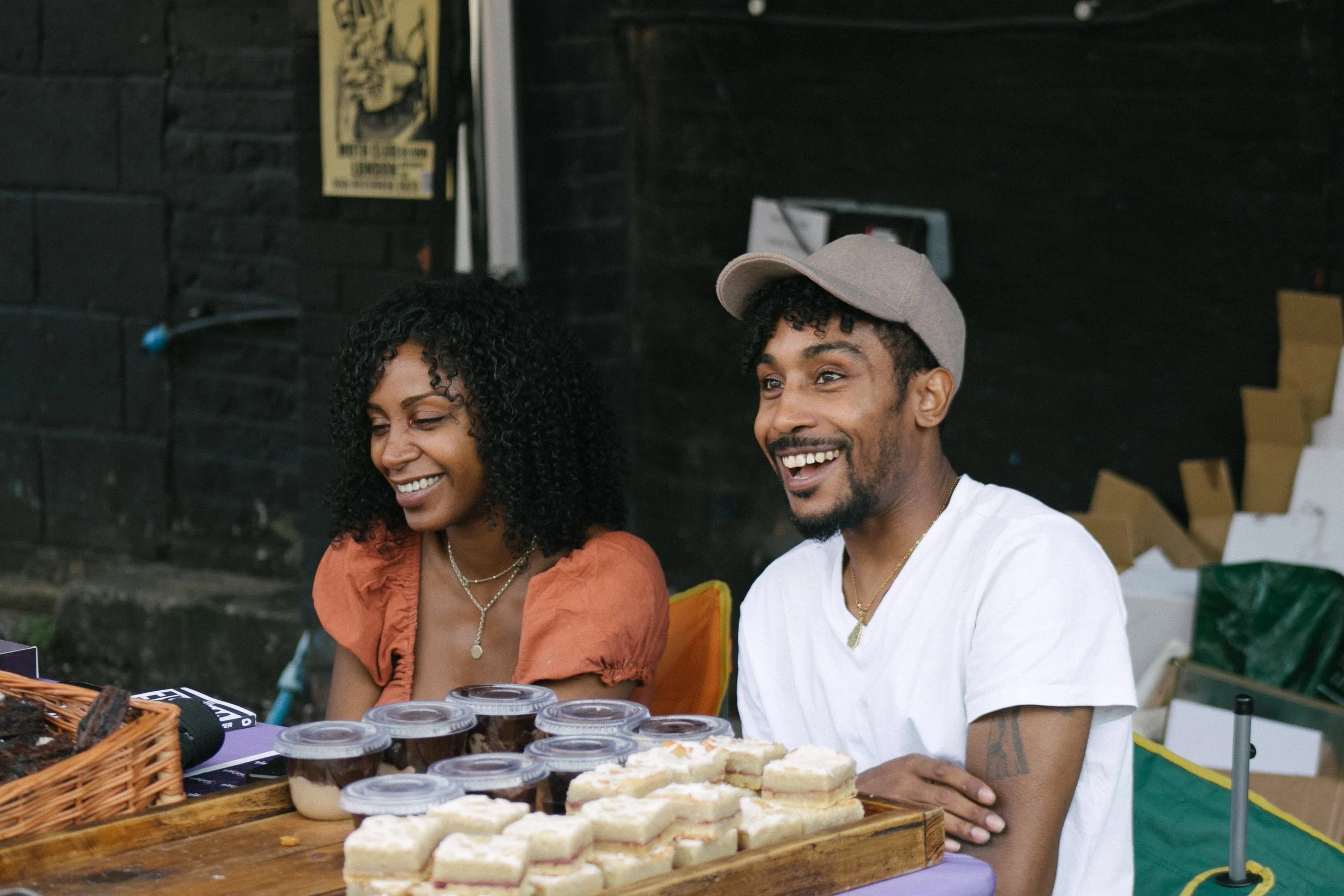 Two smiling people, a woman with curly hair and a man wearing a cap, sitting at a table with desserts and snacks.