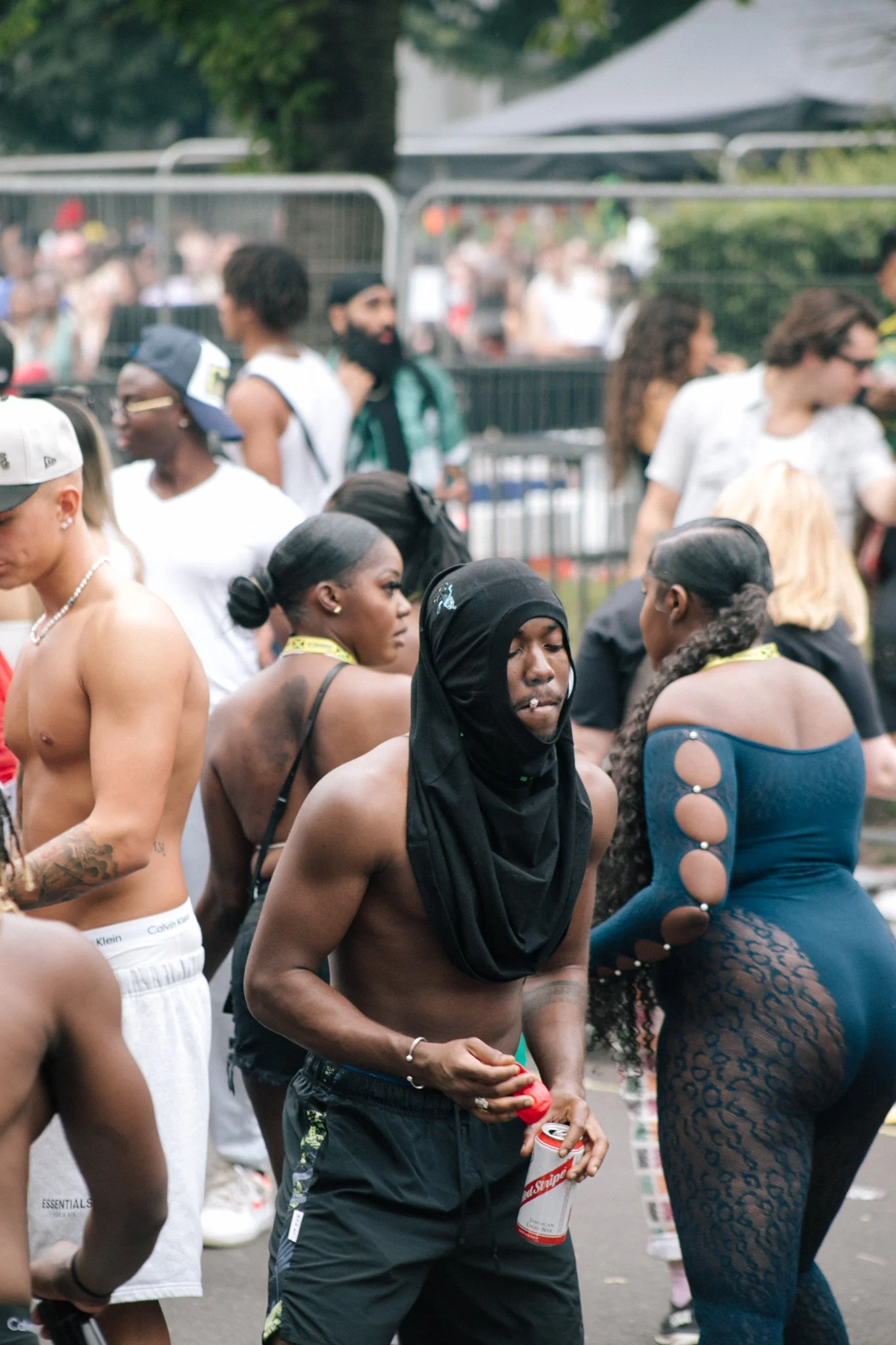 Crowd of people, some dressed in revealing clothing, at an outdoor event or festival, with barriers and trees in the background.