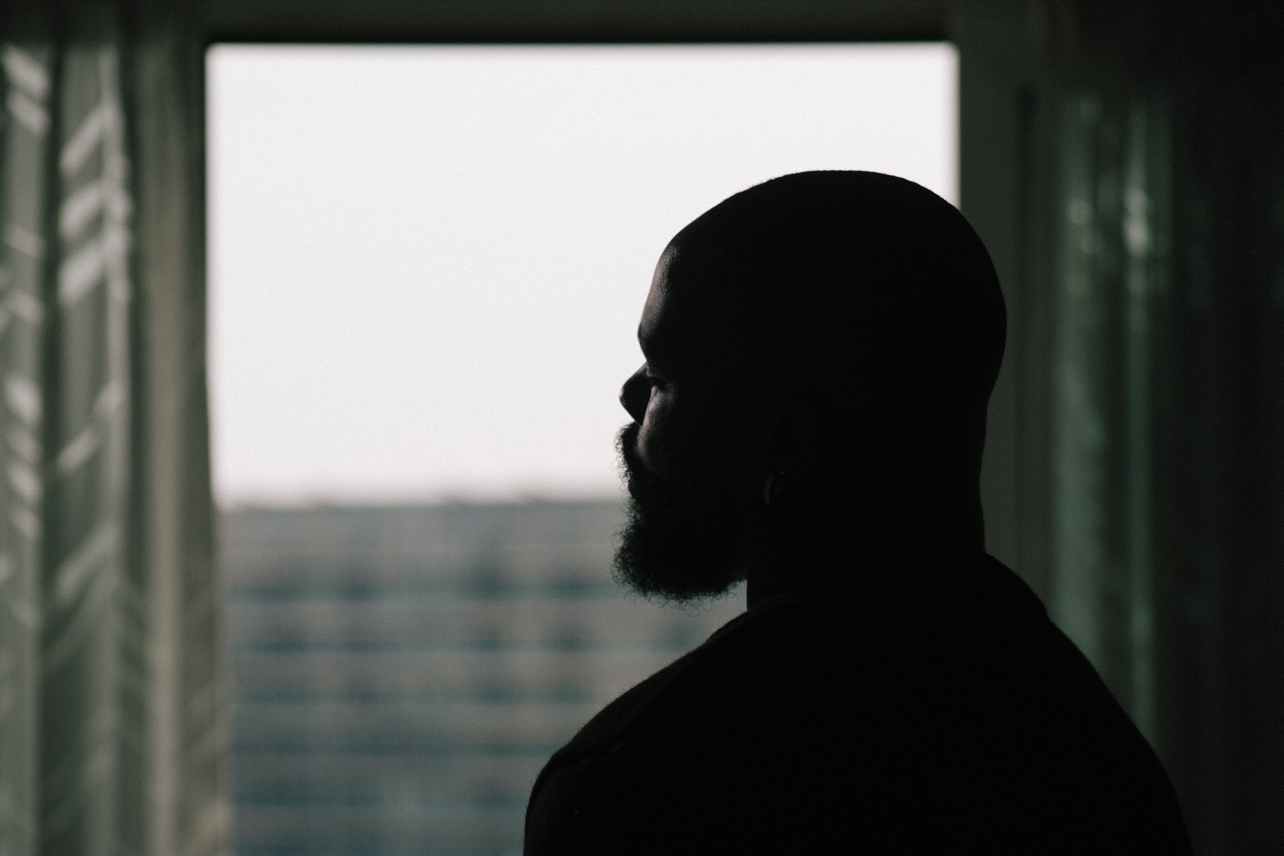 Silhouette of a man's profile with a beard, standing indoors near a window with light coming through.