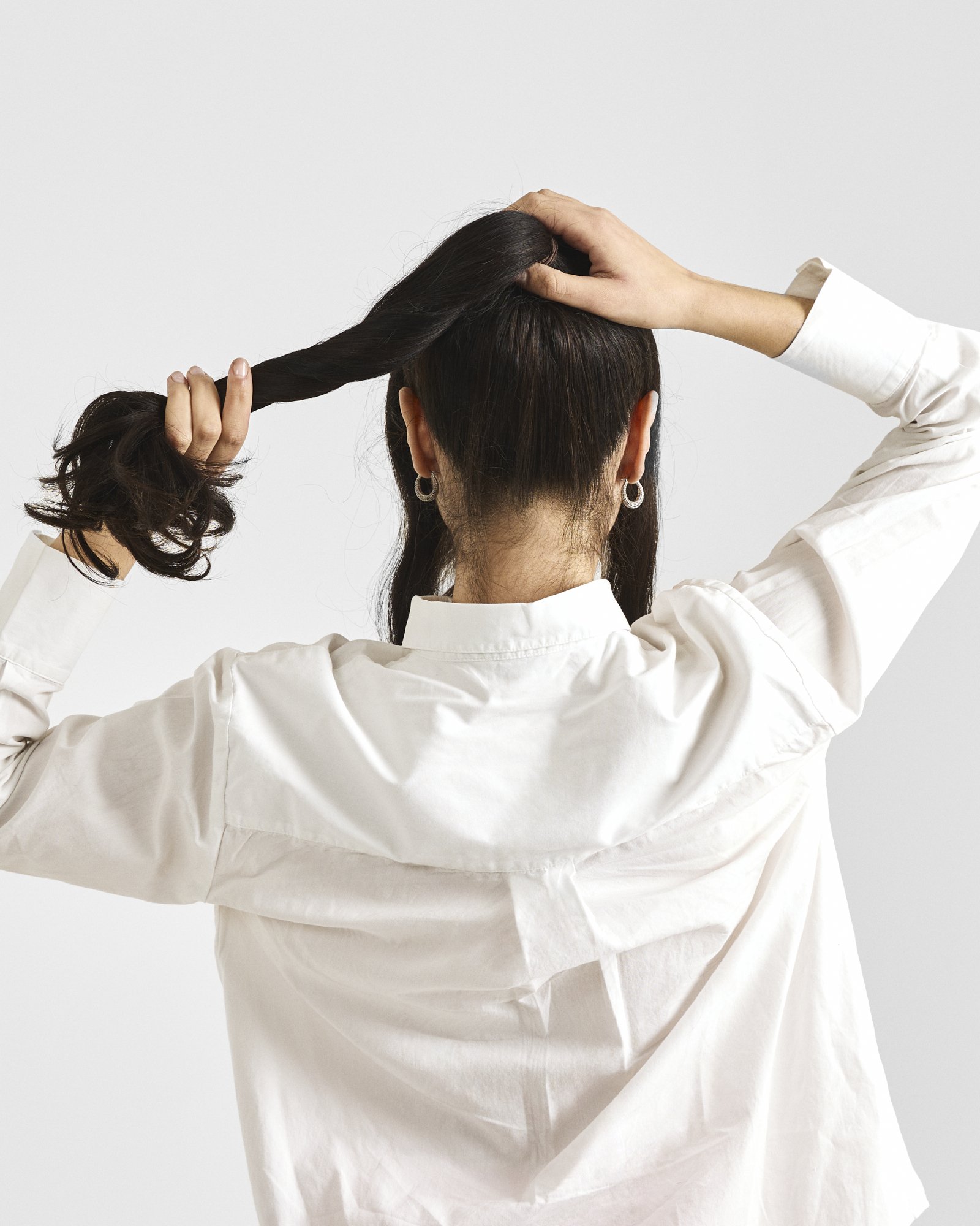 Back of a woman with dark hair wearing a white shirt, holding a twisted strand of her hair with one hand, and adjusting her hair at the top of her head with the other hand.