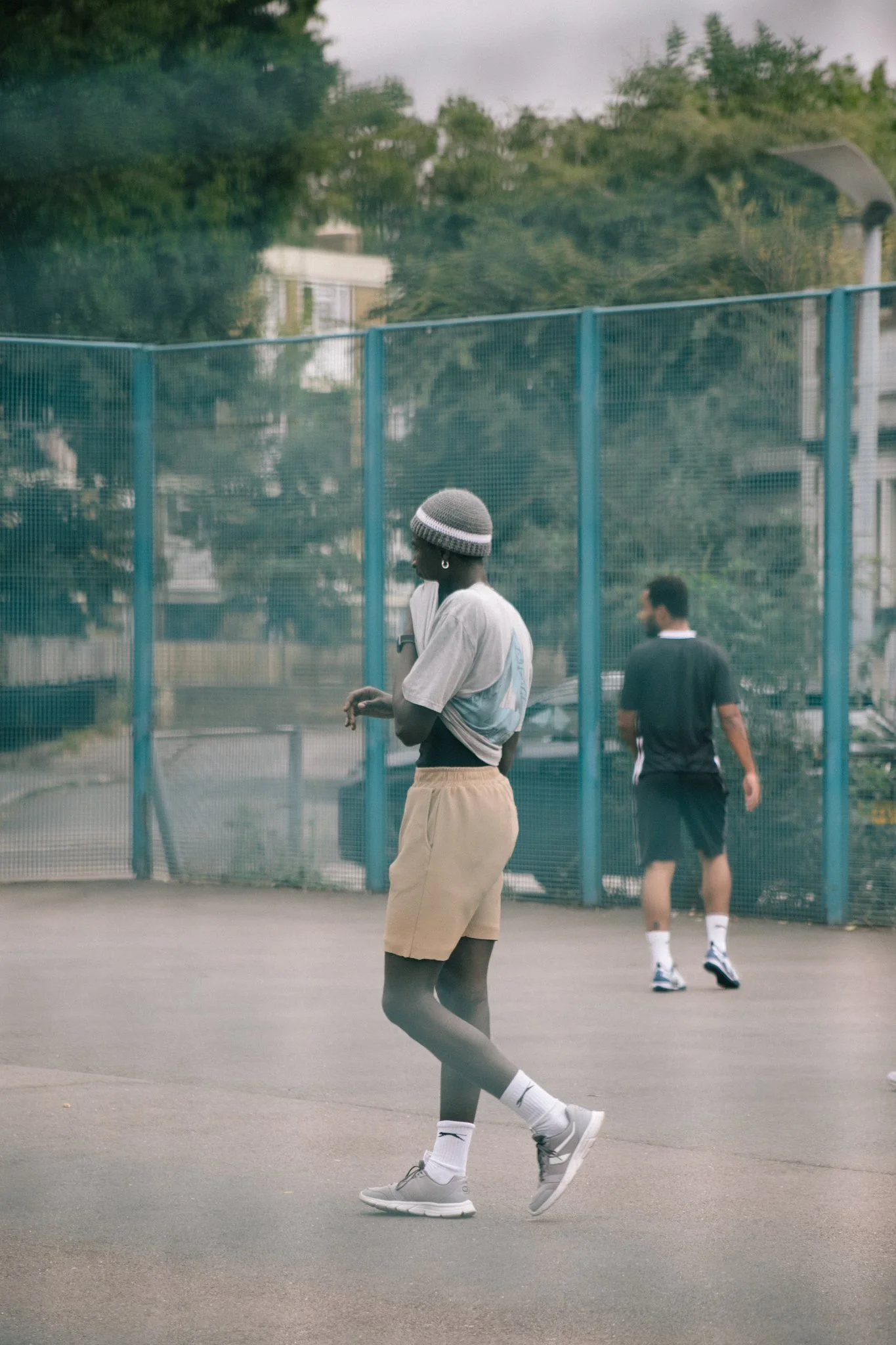 A person with dark skin wearing a gray beanie, white t-shirt, beige shorts, white socks, and gray sneakers walking on a tennis court with a safety fence. In the background, another person in black athletic shorts and a black shirt is walking, with tr