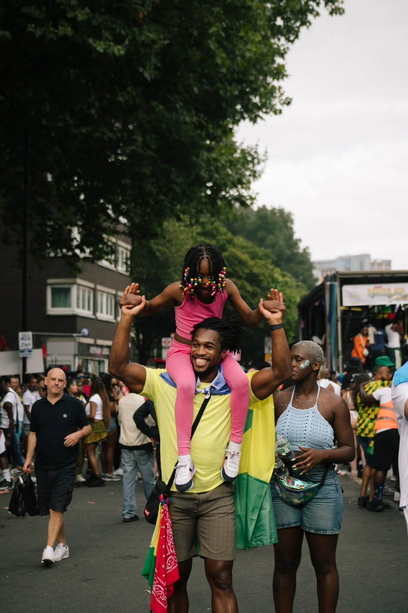 A man is smiling and carrying a young girl on his shoulders during a crowded outdoor event. The girl has colorful beads in her hair and is wearing a pink top. The surroundings show other people, some holding flags and wearing various colors, with tre