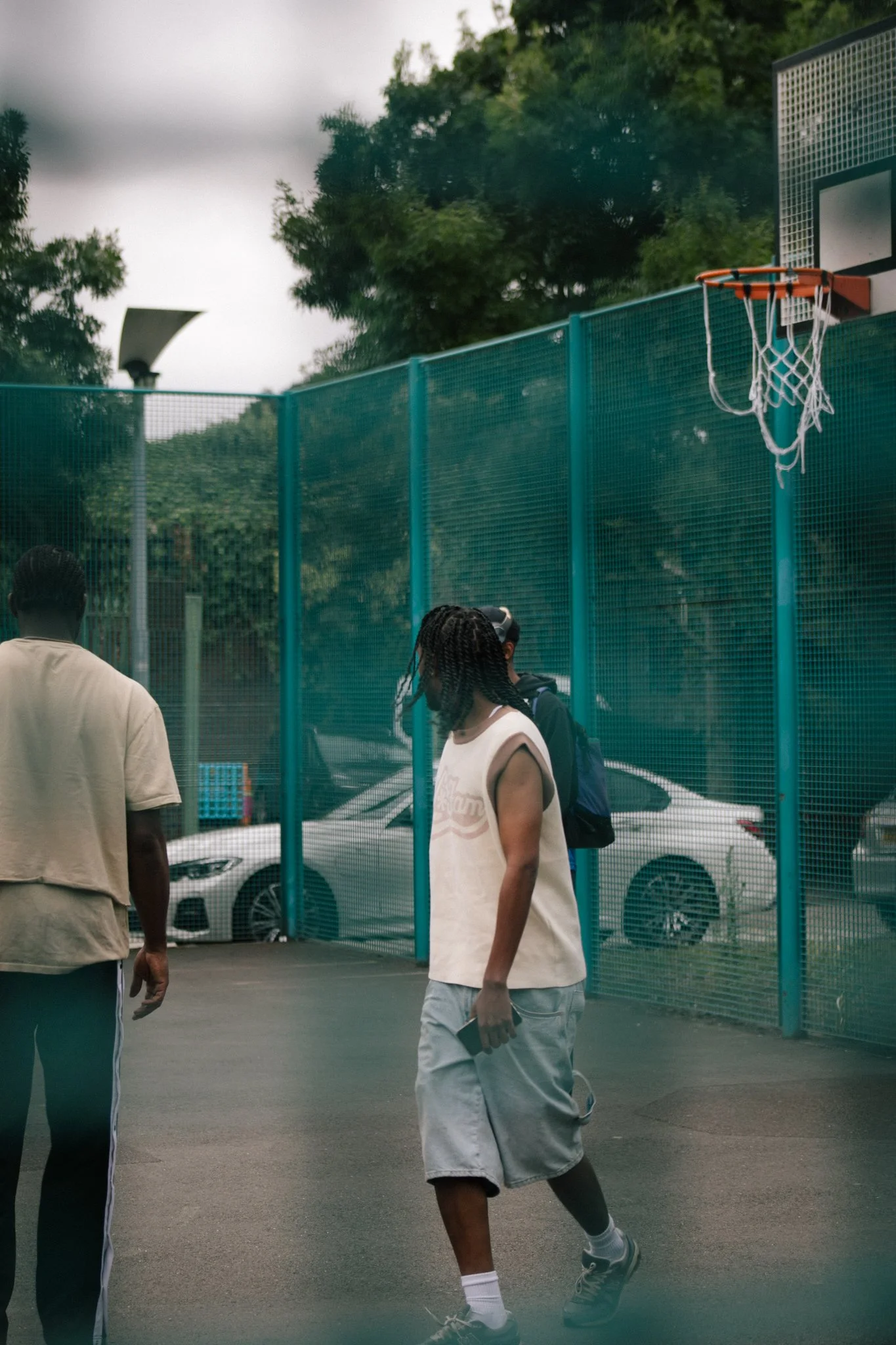 People walking in an outdoor basketball court with a hoop and a car parked nearby, surrounded by green trees.