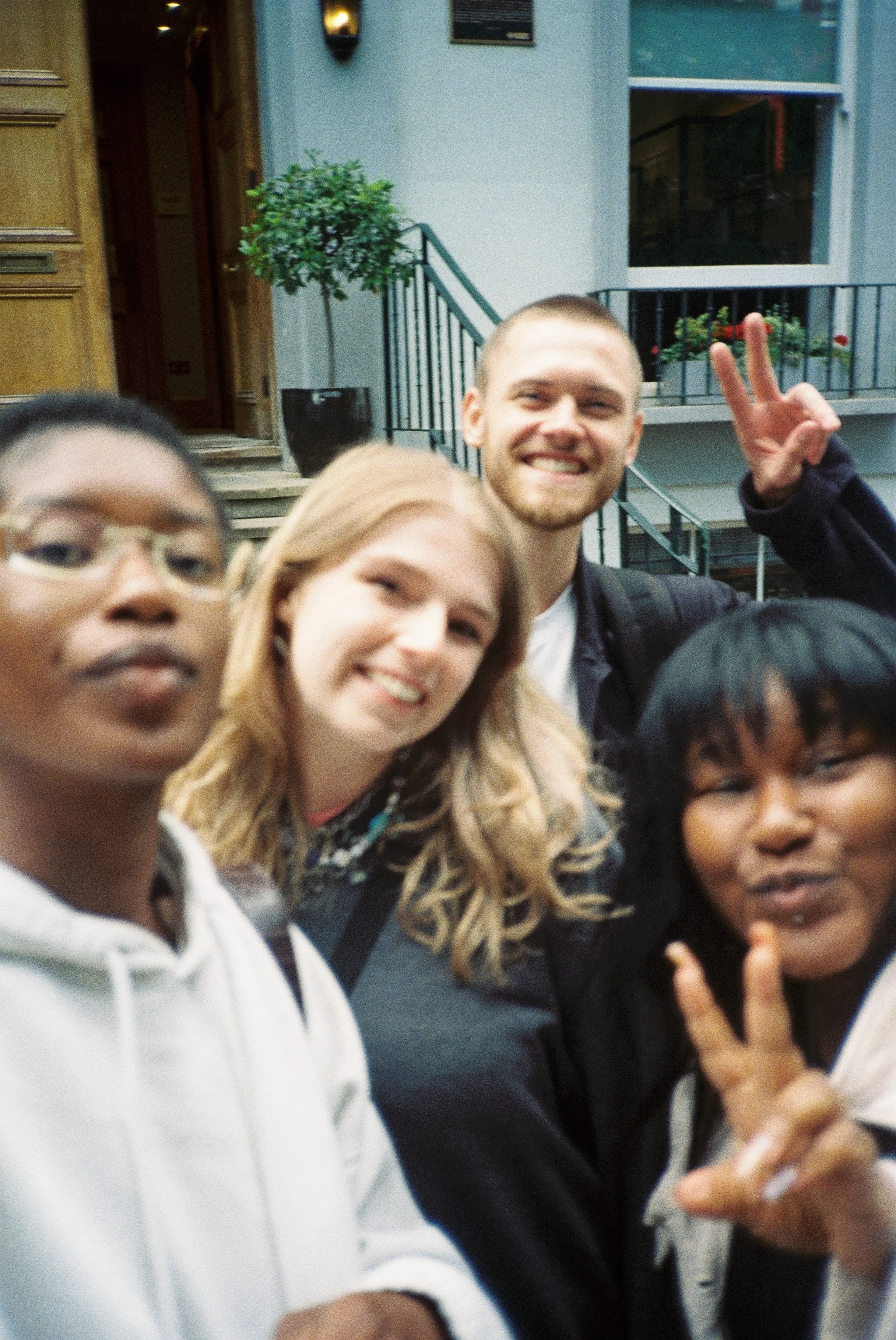 A group of young people taking a selfie outdoors, with one person flashing a peace sign, standing in front of a building entrance with plants and railings.