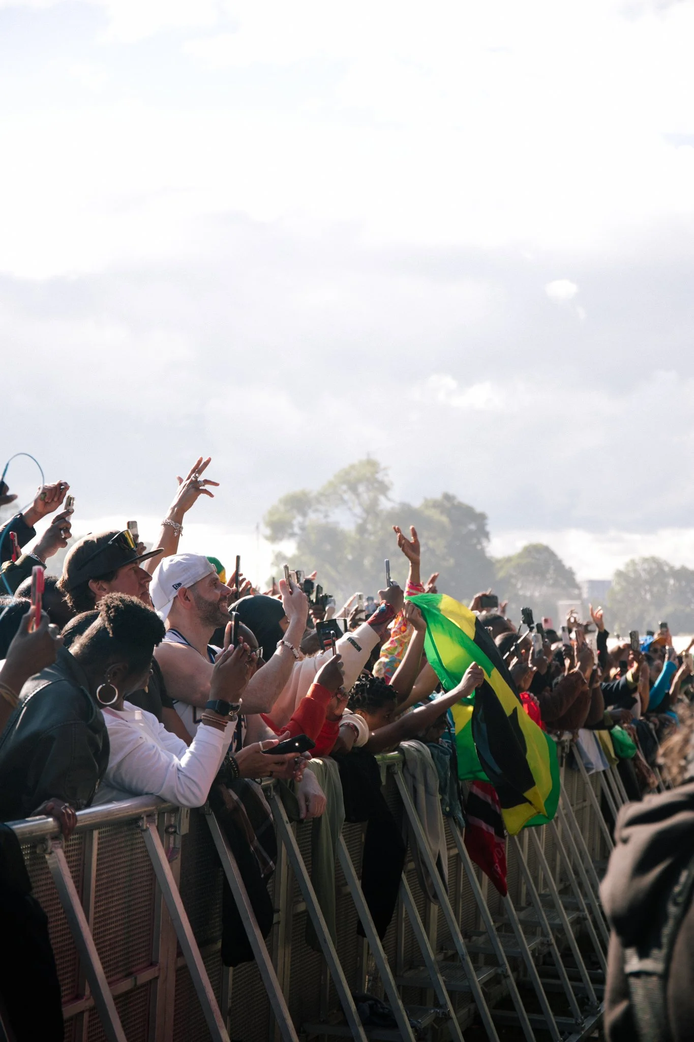 A crowd of people standing behind a metal barricade, many holding up smartphones to take pictures or videos. Some individuals are waving or holding flags, with a partly cloudy sky and trees in the background.