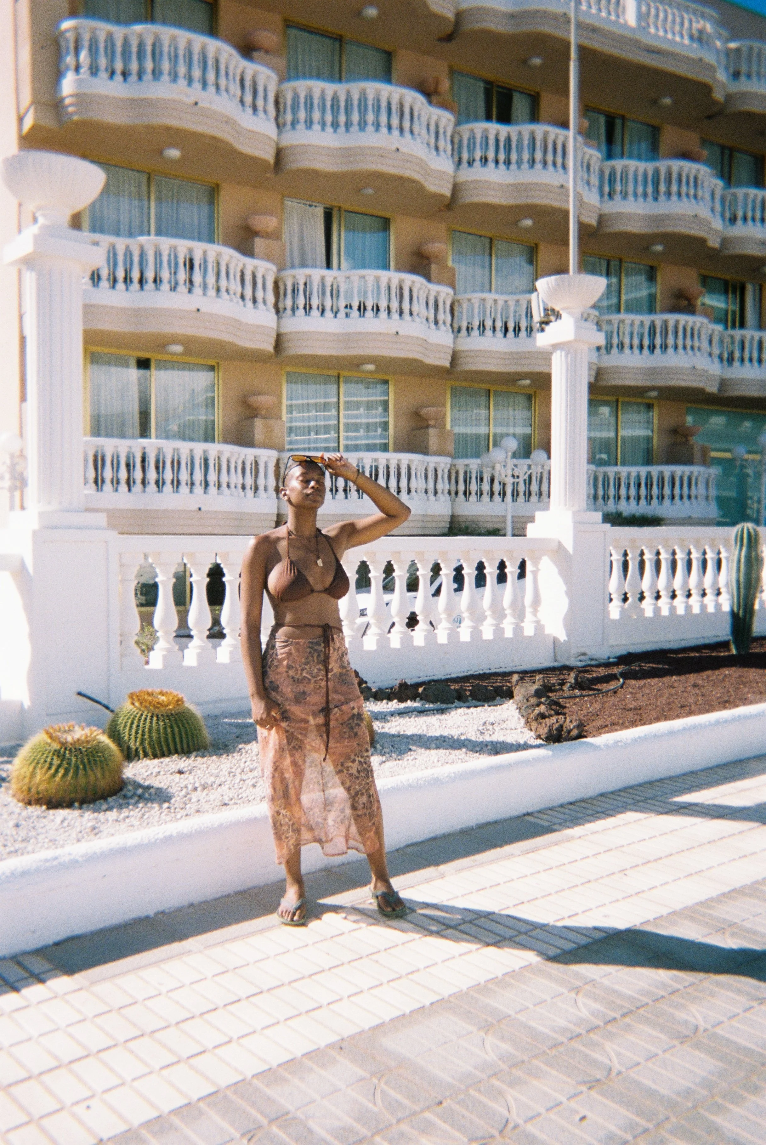 A woman in a brown bikini top and patterned skirt holds her sunglasses while standing in front of a hotel with multiple balconies and white fence. There are cacti and a white stone border in the foreground.