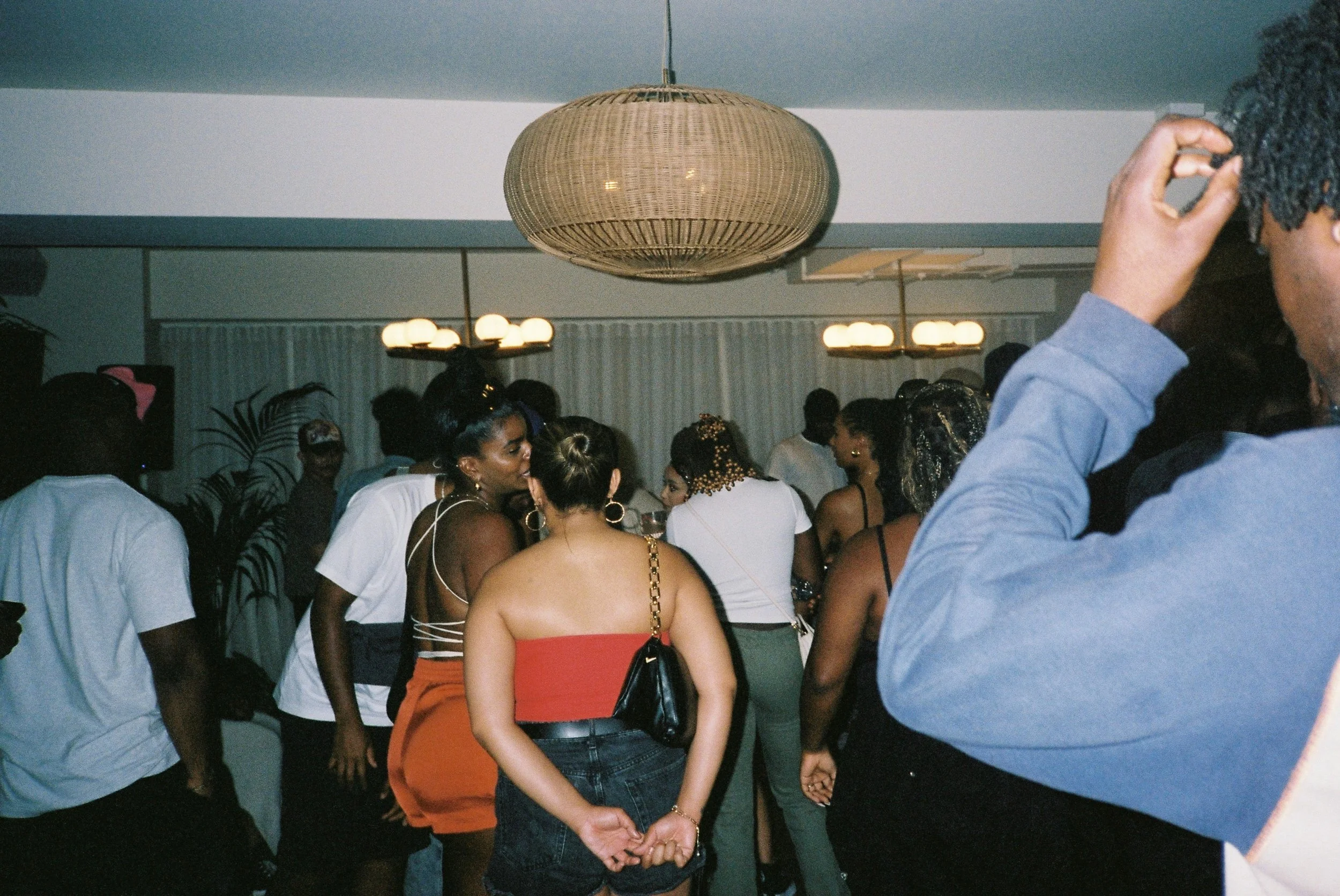 A crowded indoor event with people socializing and dancing, illuminated by warm lighting fixtures, including a large woven pendant lamp.