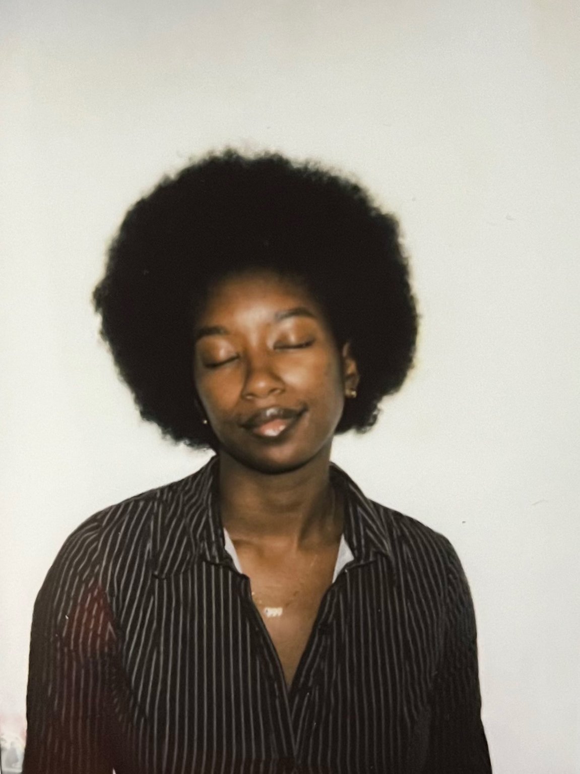 A woman with closed eyes, an afro hairstyle, wearing a brown and white striped shirt, standing against a plain white background.