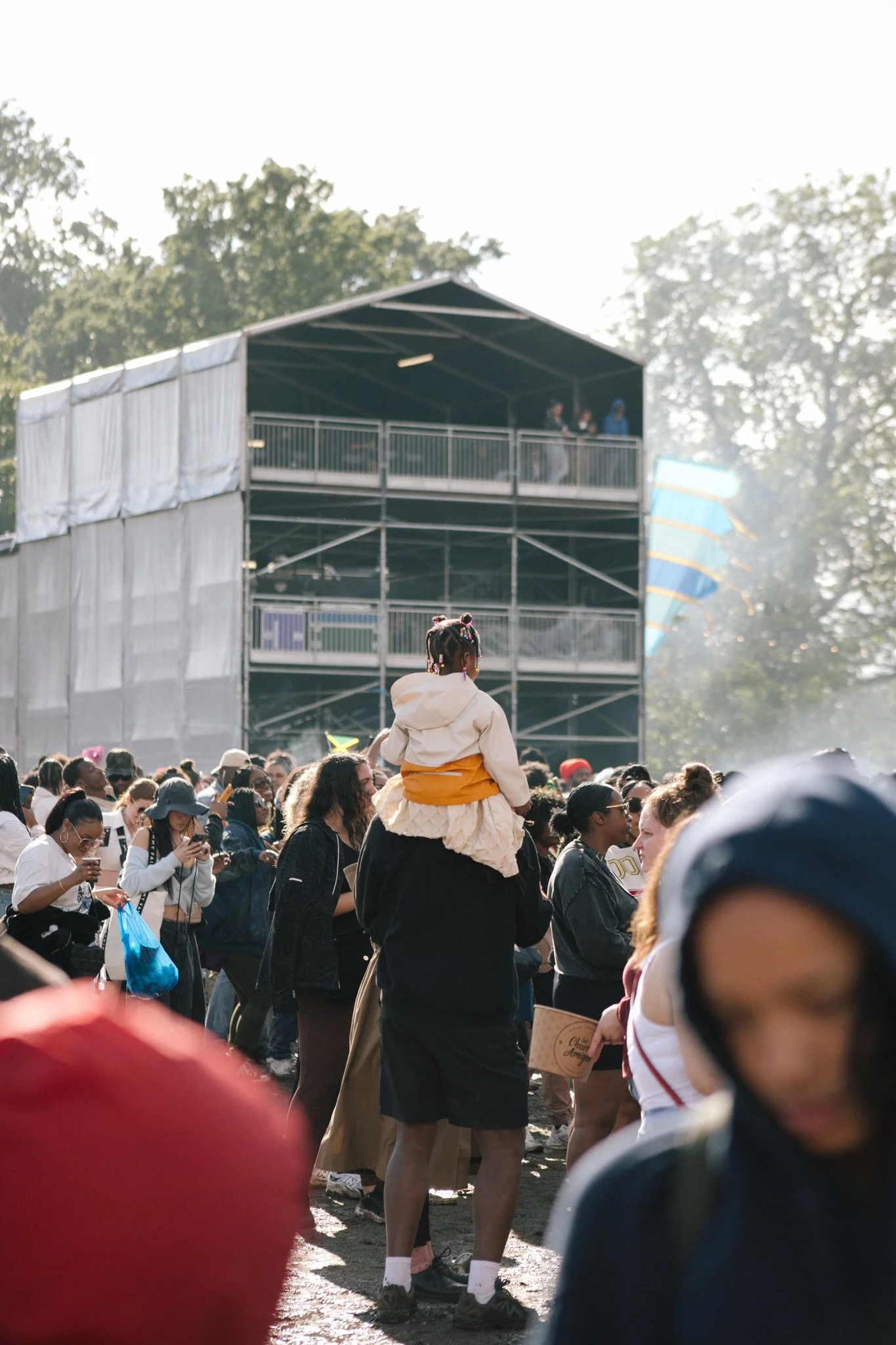 Crowd of people gathering outdoors with a person on someone's shoulders, a stage or scaffolding structure in the background, and trees; some people are using their phones.