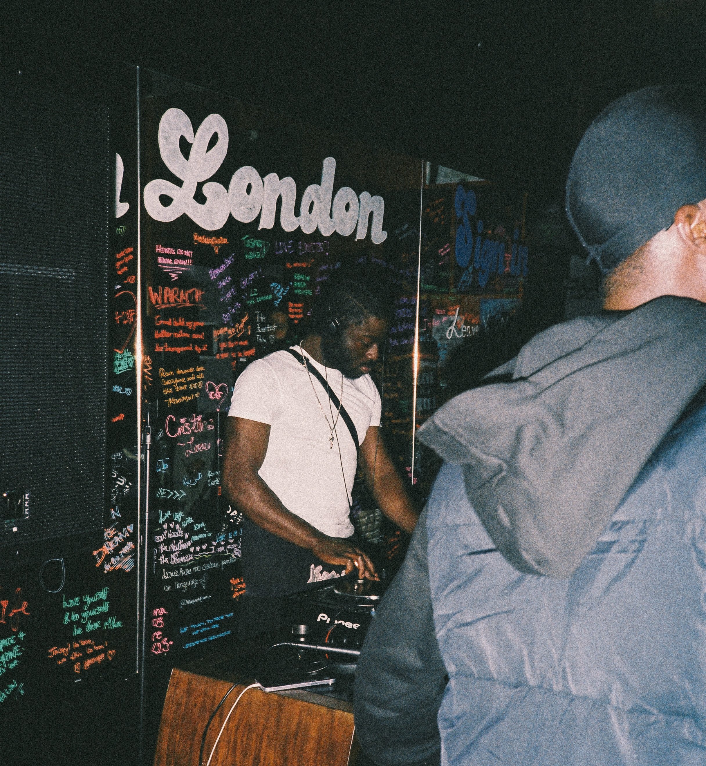 People at a DJ booth with colorful chalkboard signs in the background, including the word "London."