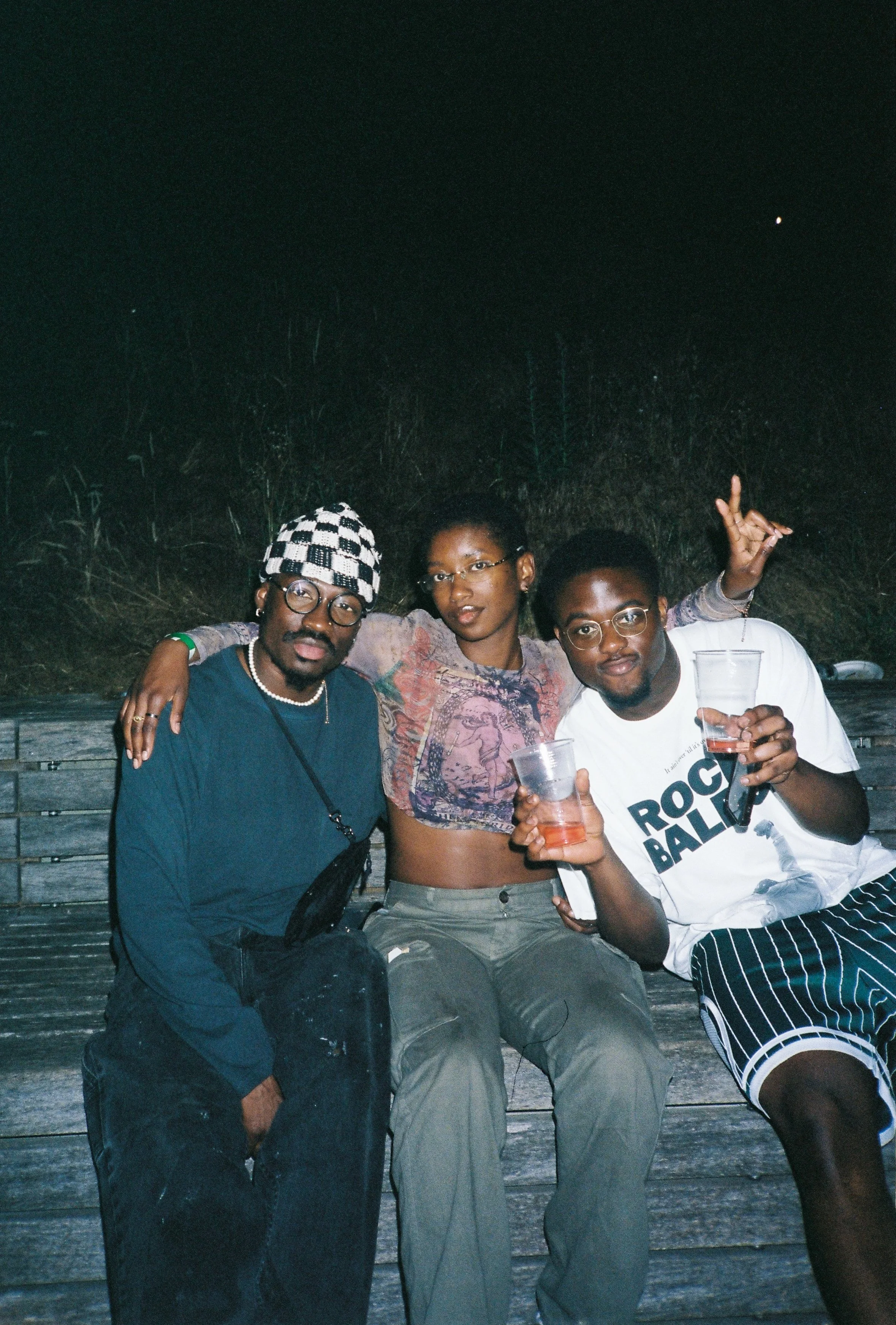 Three friends sitting together on a wooden bench outdoors at night, smiling and holding drinks, with a dark sky in the background.