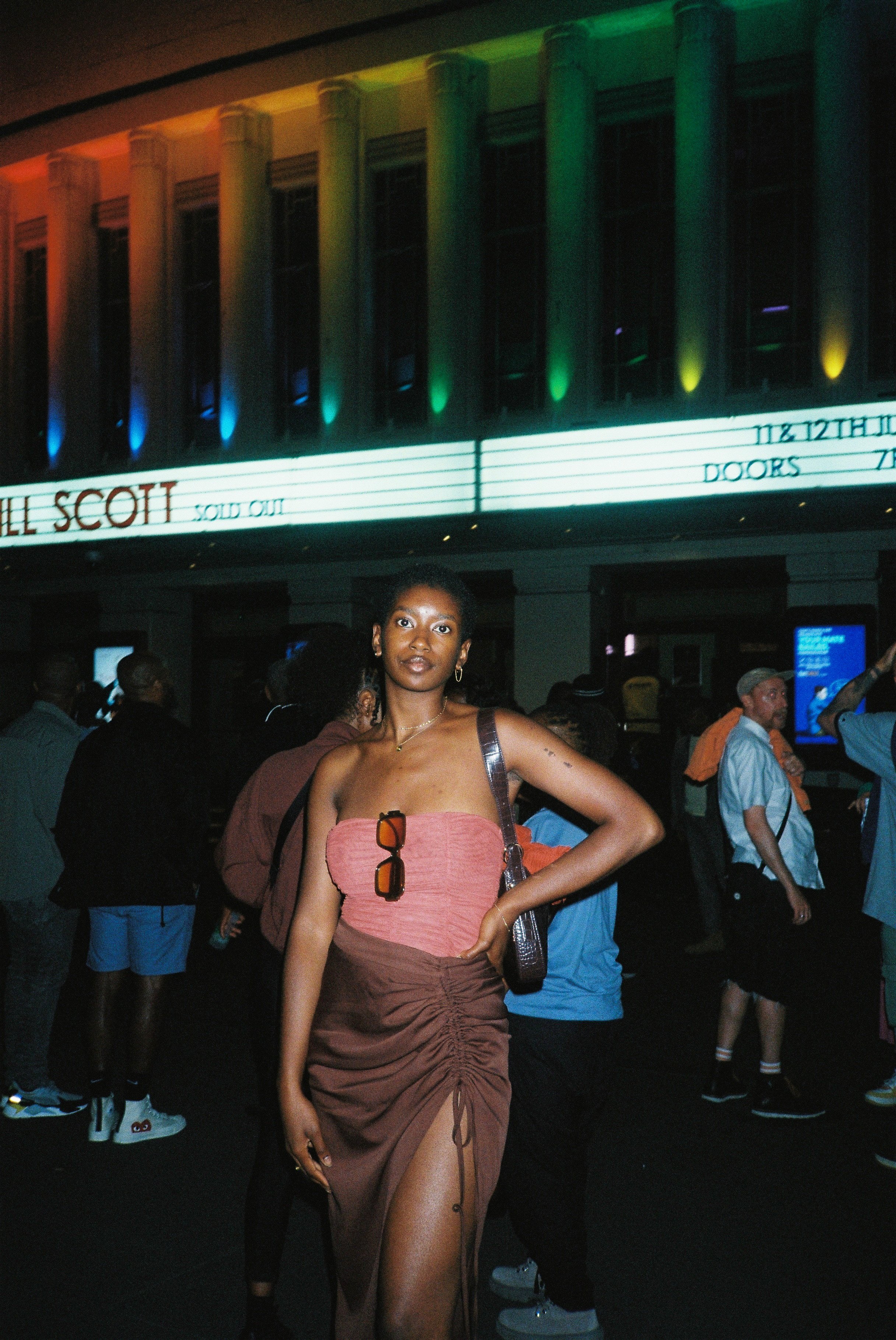 A woman posing confidently with one hand on her hip in a crowded area at night, in front of a building featuring colourful lights in the background and a sign that reads Jill Scott