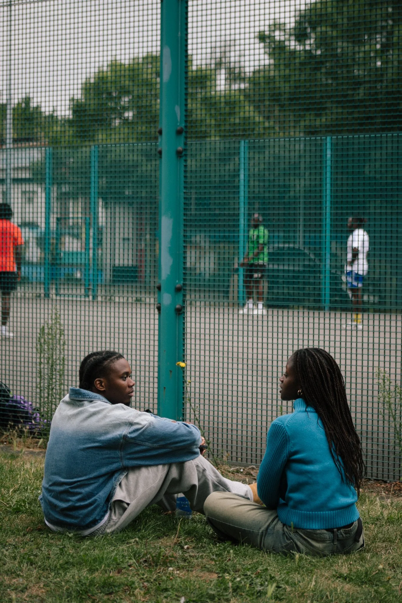 Two people sitting on grass in front of a green fence, having a conversation. In the background, men are playing footbal behind the fence, one in a green shirt and the other in a white shirt.