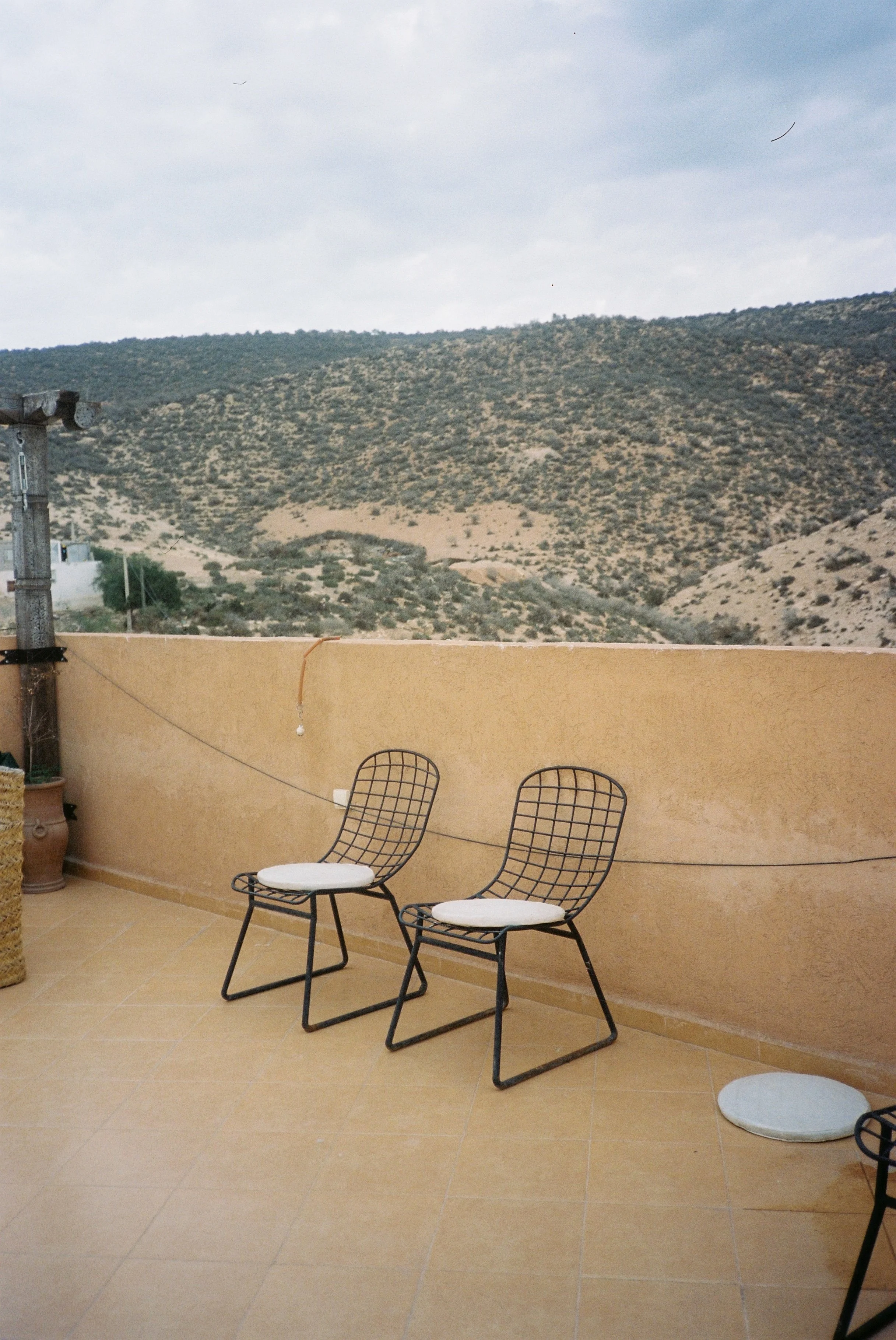 A patio with two black metal chairs with white cushions, a yellow wall, potted plant, and a mountainous landscape with scattered shrubs in the background under a cloudy sky.