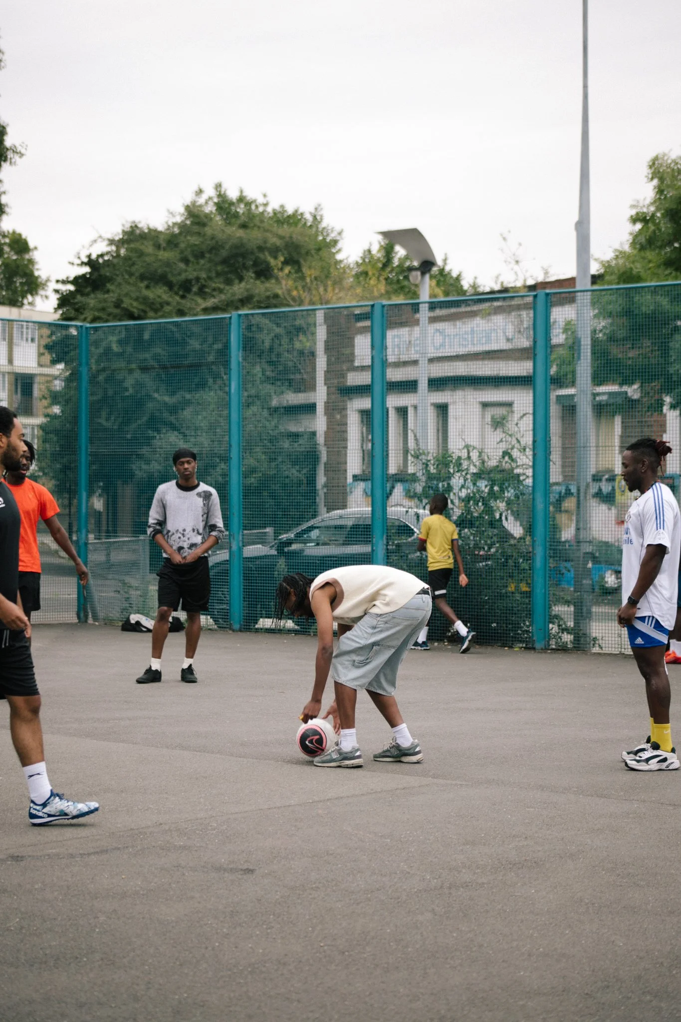 A group of young men playing football on a court enclosed with blue fencing. One person is bending down to place a black and pink football. Several others are standing around, and there are trees and a building in the background.