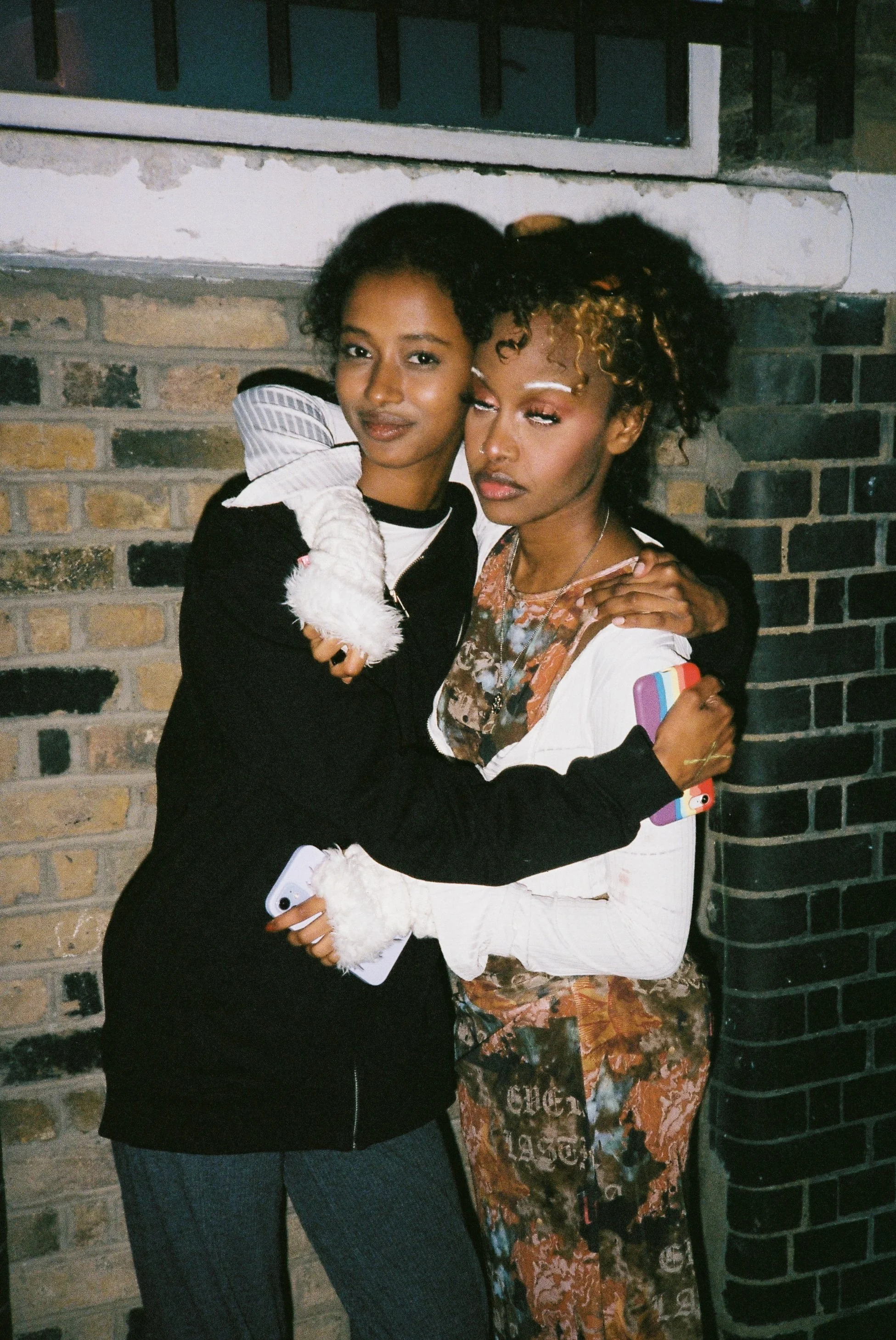 Two women hugging each other in front of a brick wall. One woman has dark hair and is wearing a black jacket, the other woman has curly hair with blonde highlights and is wearing a white jacket with patterned dress underneath.