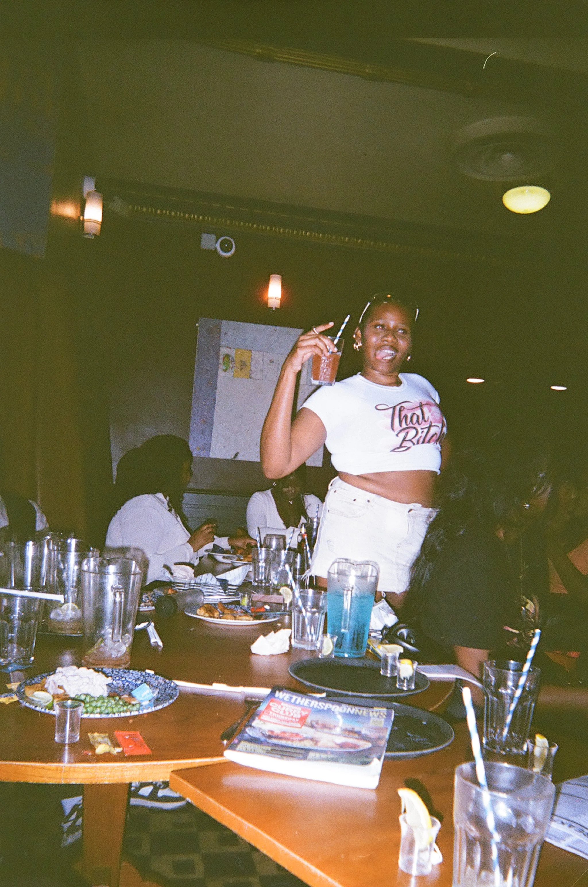 A woman standing in a bar, holding a drink and smiling. She is wearing a white crop top. The table in front of her has plates with food, drinks, and utensils, and there are other people seated around the table under dim lighting.
