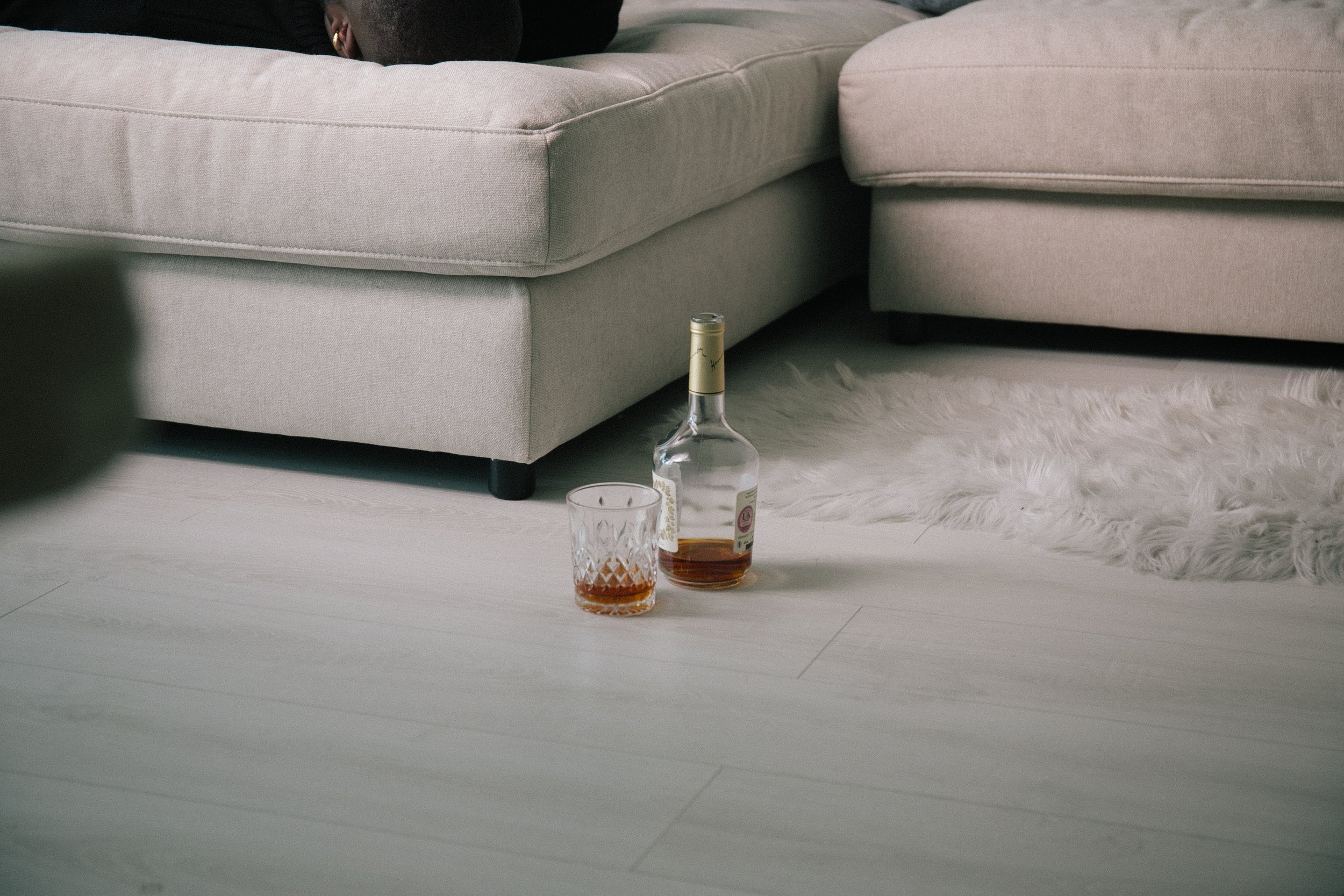 A beige sofa with a person lying on it, partially visible. On the floor in front of the sofa, there is a bottle of Hennesy and a glass with some liquid, placed on a fluffy white rug.