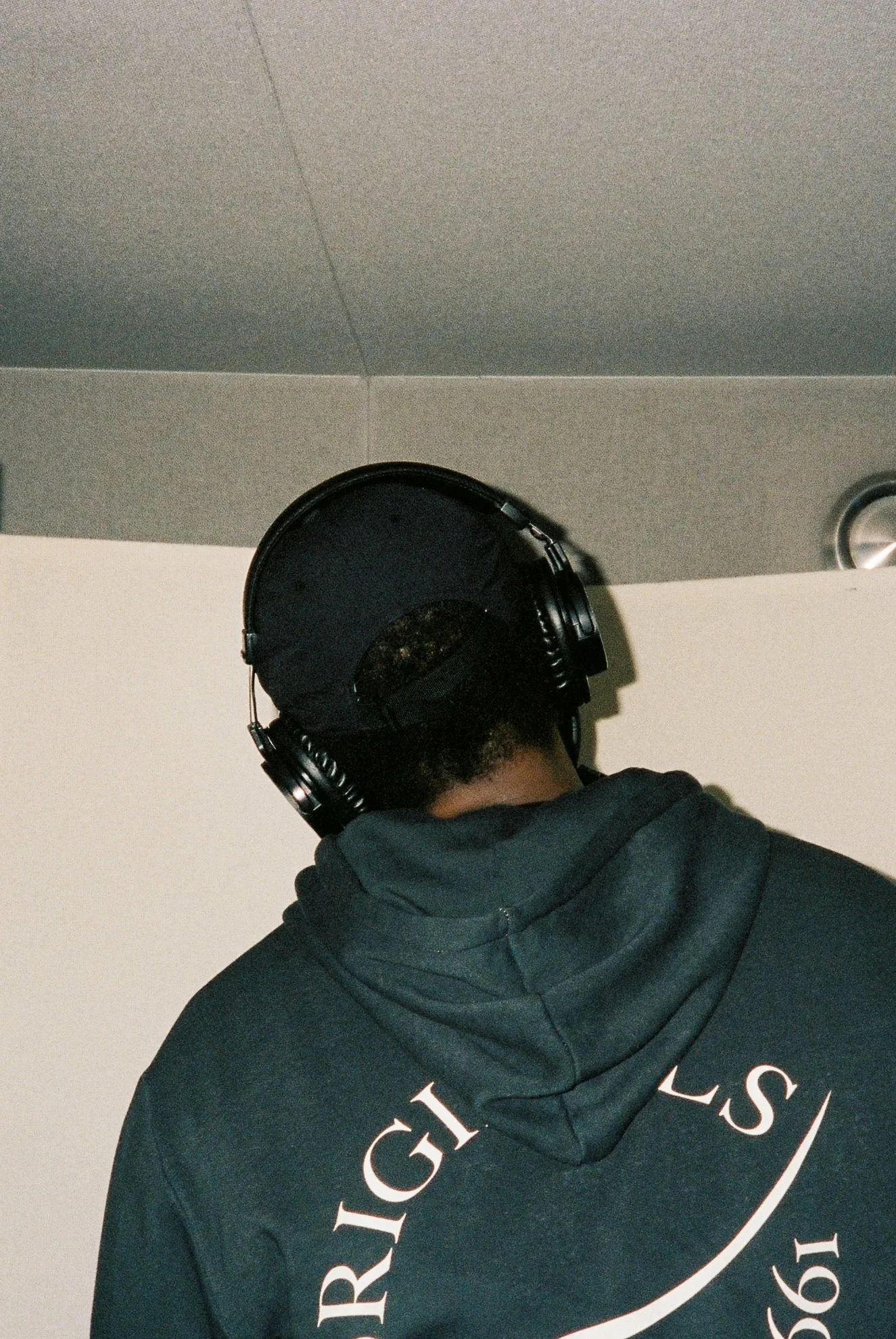 Person wearing a dark hoodie with white text on the back, black headphones, and standing in a studio setting.