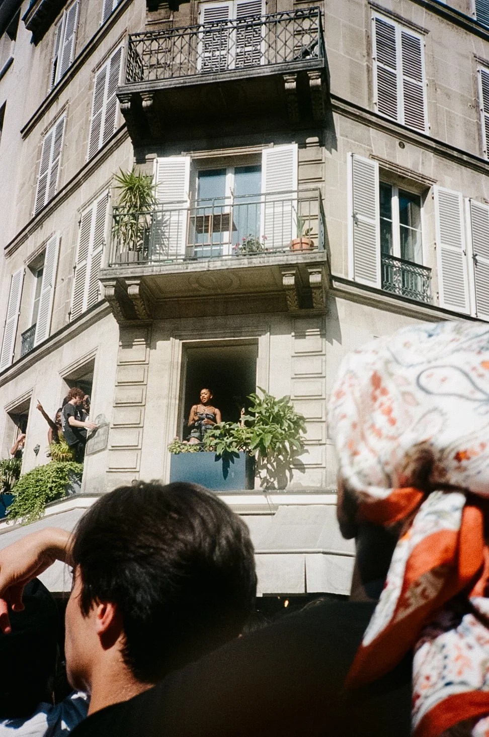 People gathered outside and looking up at a similar building with balconies, some with plants, in a city.