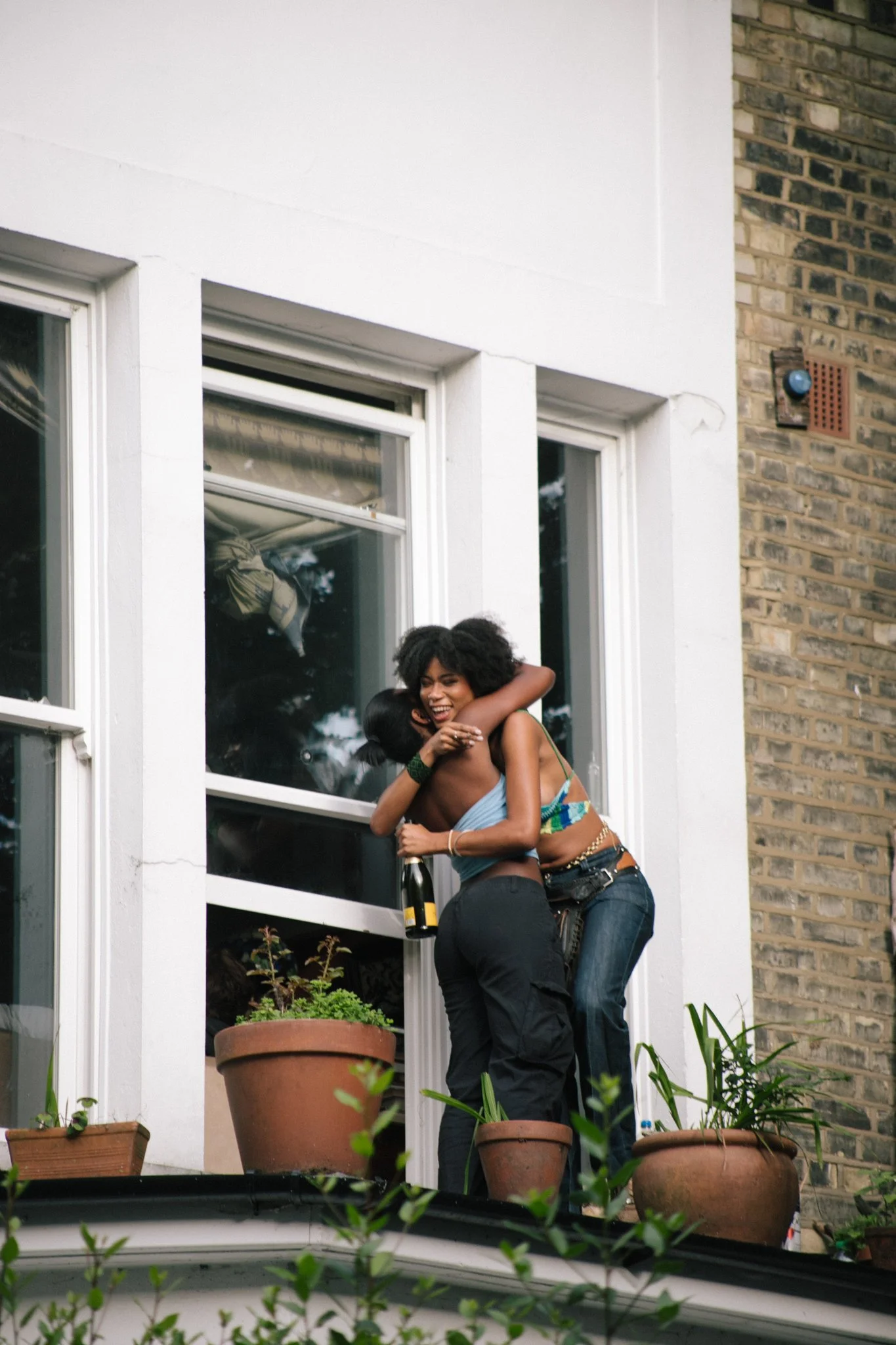 Two women hugging and smiling on a balcony outside a house with windows and potted plants.