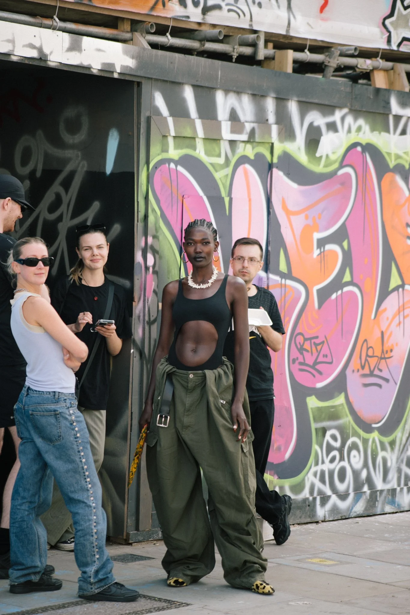A diverse group of people standing and leaning against a graffiti-covered wall on a city sidewalk, with one person dressed in baggy pants and a black tank top, wearing leopard print shoes, and others casually dressed with sunglasses and backpacks.
