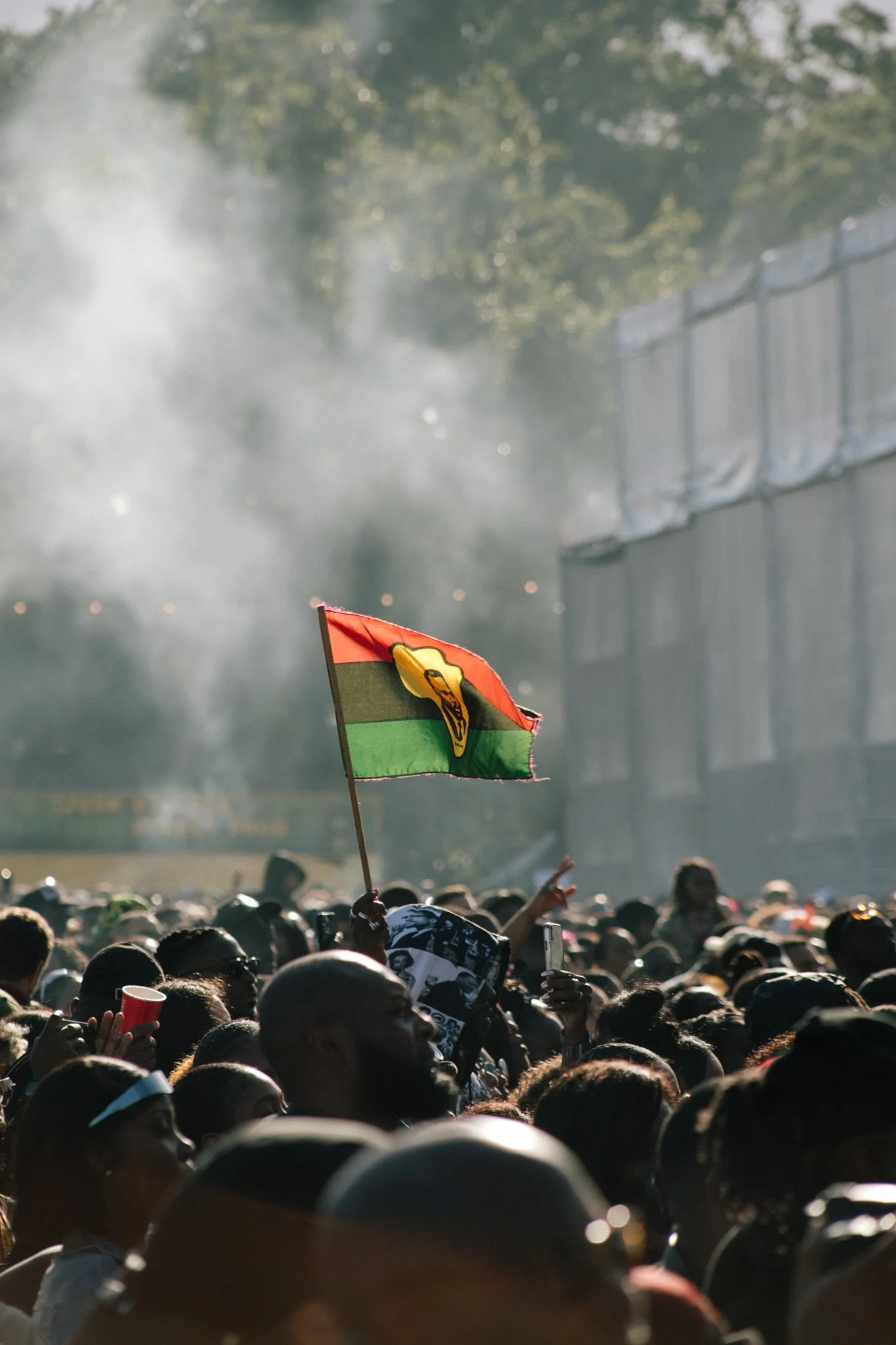 A large crowd at an outdoor event, with a small colorful flag being waved among the attendees in the foreground. Smoke and trees are visible in the background.