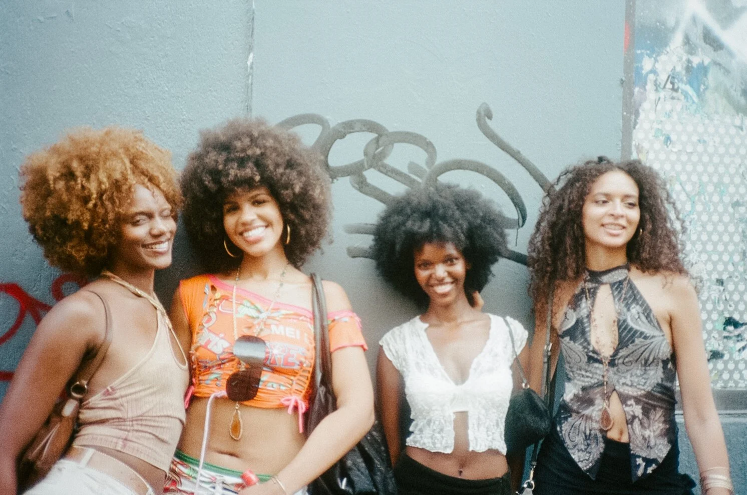 Four young women with curly hair smiling in front of a wall with graffiti of interlocked rings.
