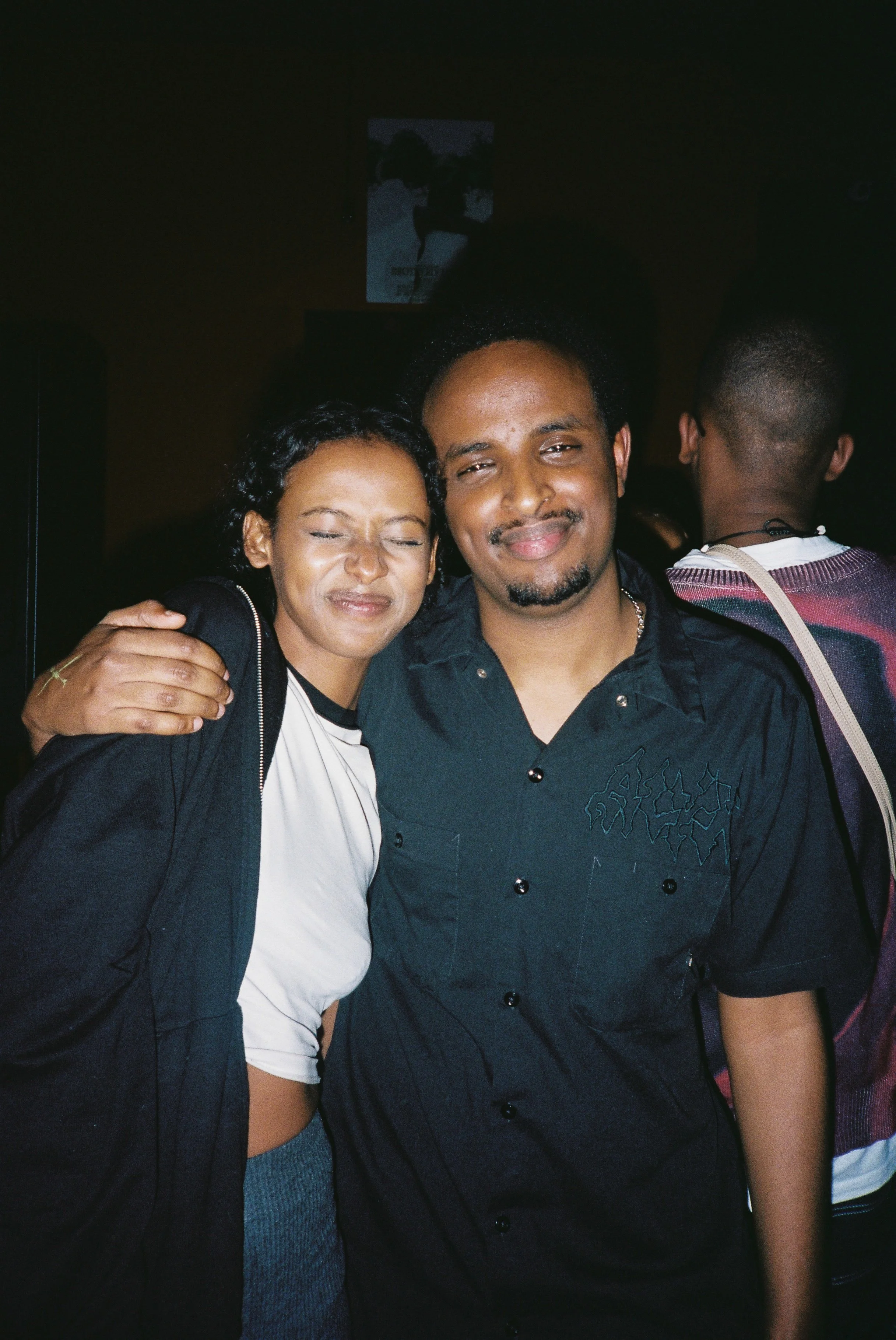 A woman with curly black hair and light brown skin smiling with her eyes closed, hugging a man with light brown skin and a short beard, who is also smiling. The setting appears to be a crowded indoor space, possibly a party or event.
