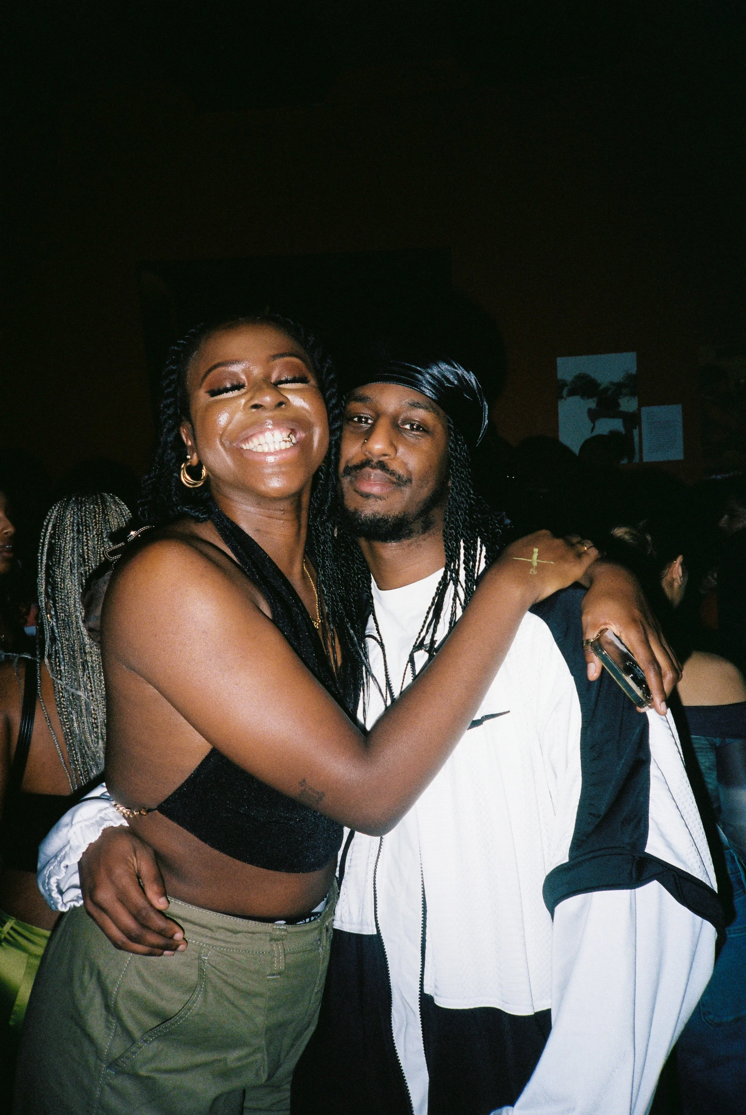 A smiling woman with dark curly hair, wearing a black sleeveless top and green cargo pants, embracing a man with their hair in twists, a black headscarf, and a nose piercing, in a dimly lit setting.