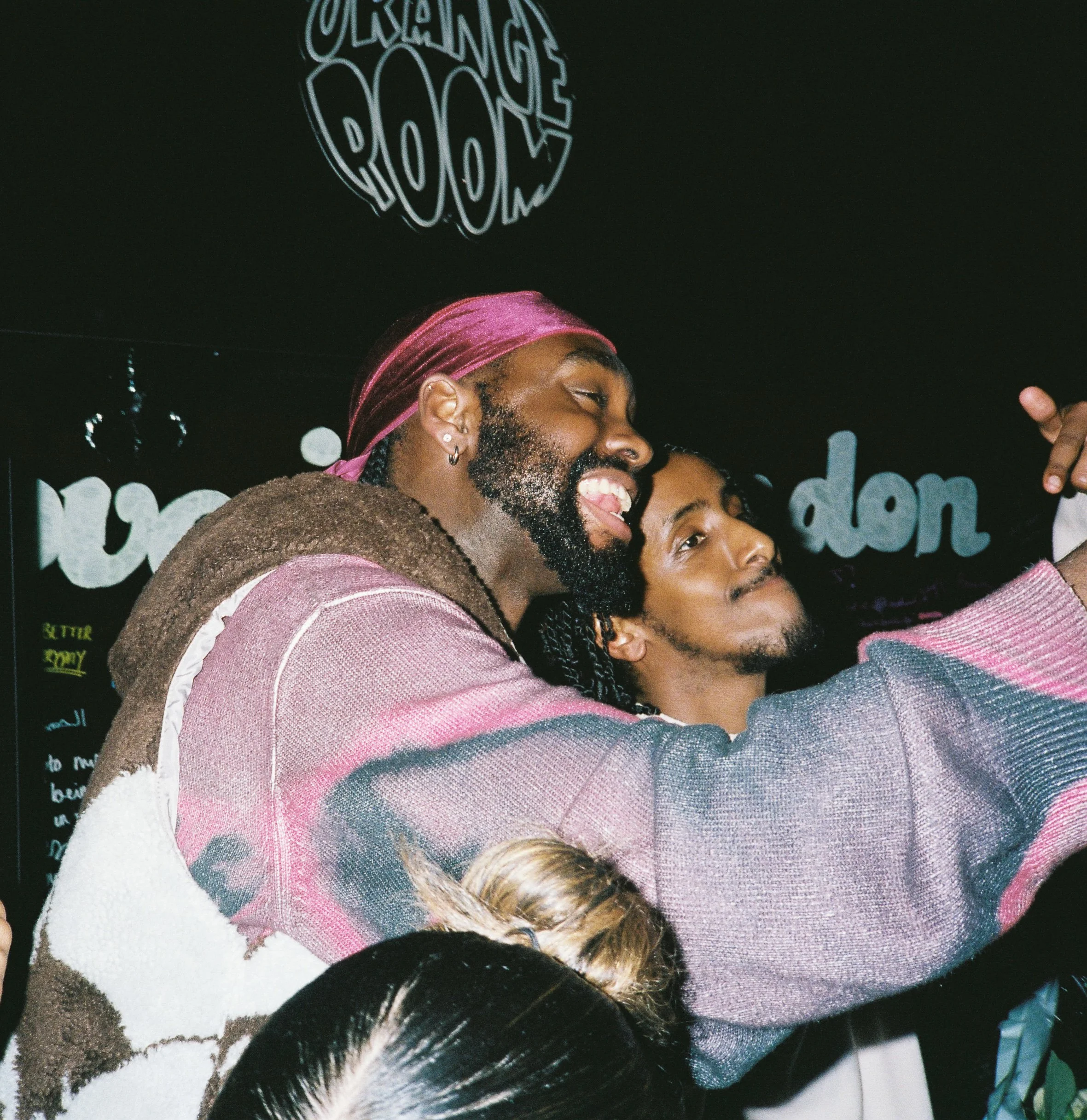 Two people are embracing and smiling, wearing casual clothing and posing for another photo. One person is wearing a pink durag and has earrings, while the other has short curly hair.
