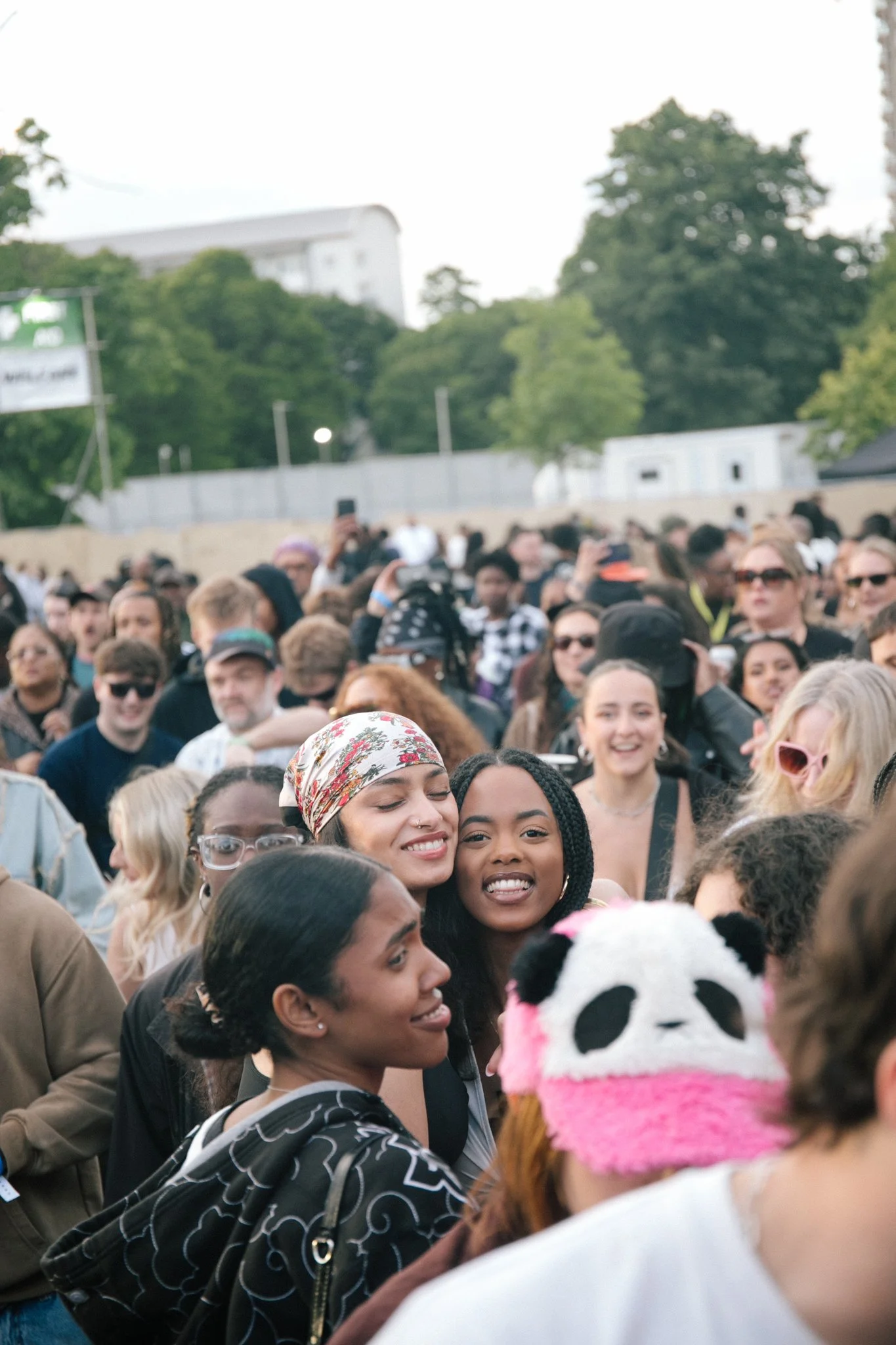 Crowd of diverse people smiling and socializing outdoors, with trees and buildings in the background.