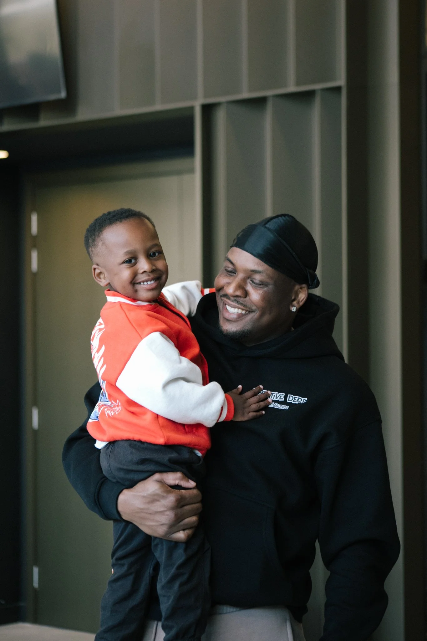 A young black boy with a big smile being held by an adult black man with a big smile in an indoor setting with gray walls.