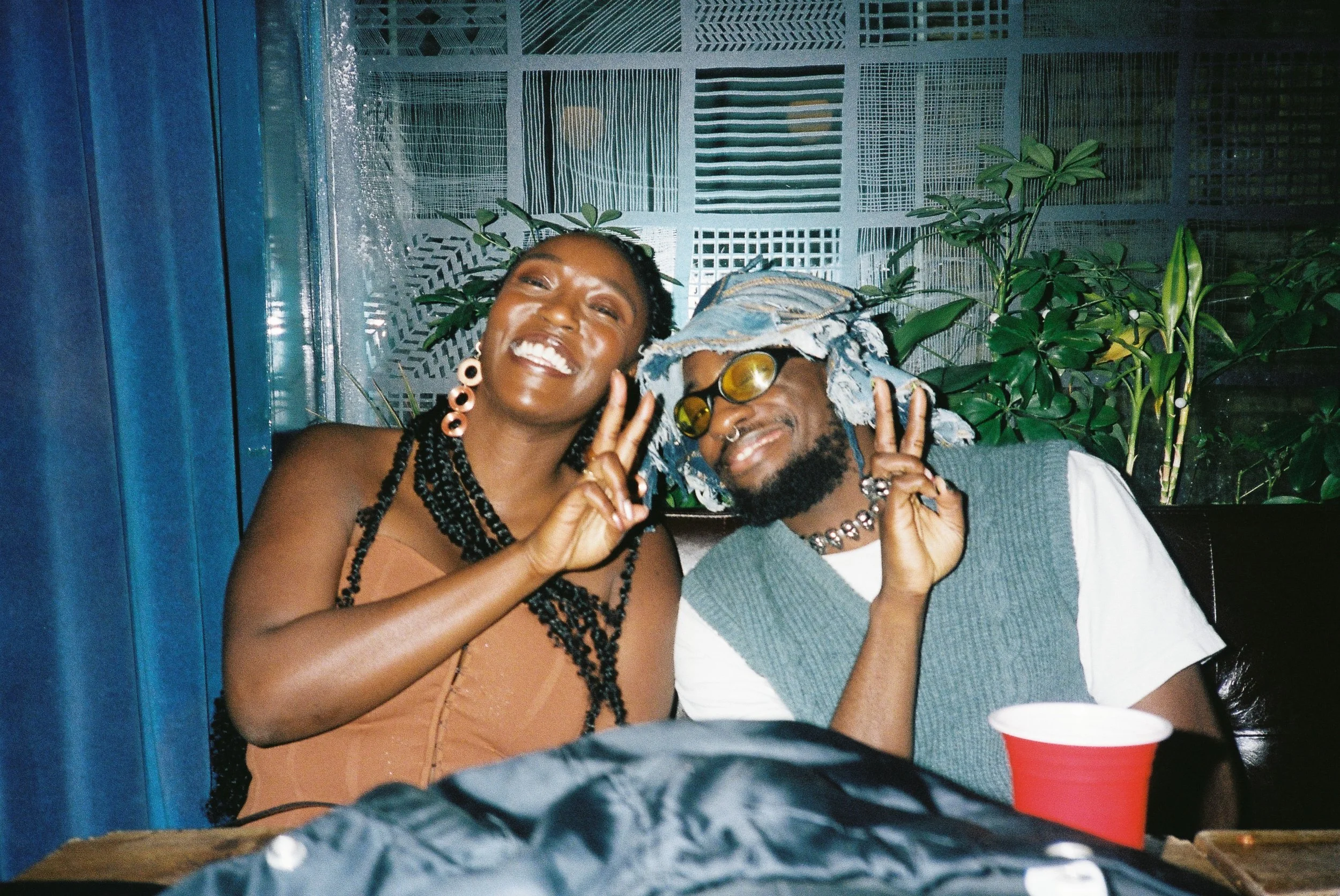 Two smiling people, a woman with braids and large earrings and a man with glasses and a bucket hat, posing with peace signs at a table with a red cup, in a setting with plants and patterned wall panels in the background.