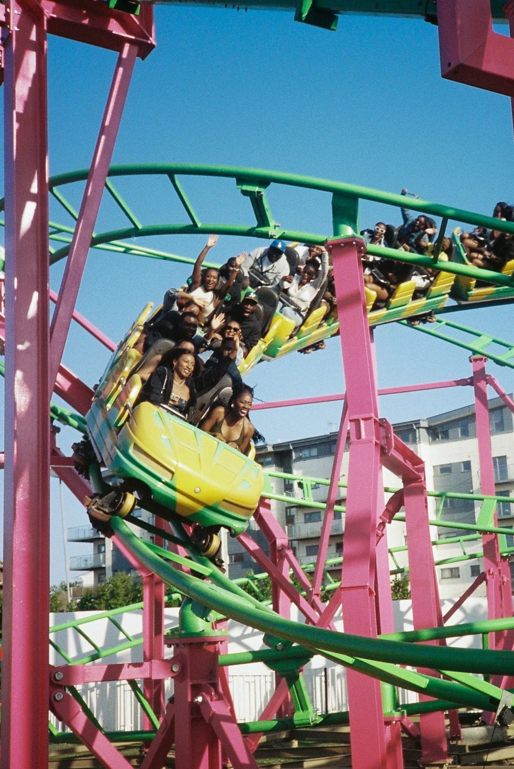 People riding a colorful roller coaster at an amusement park on a sunny day, smiling and waving.