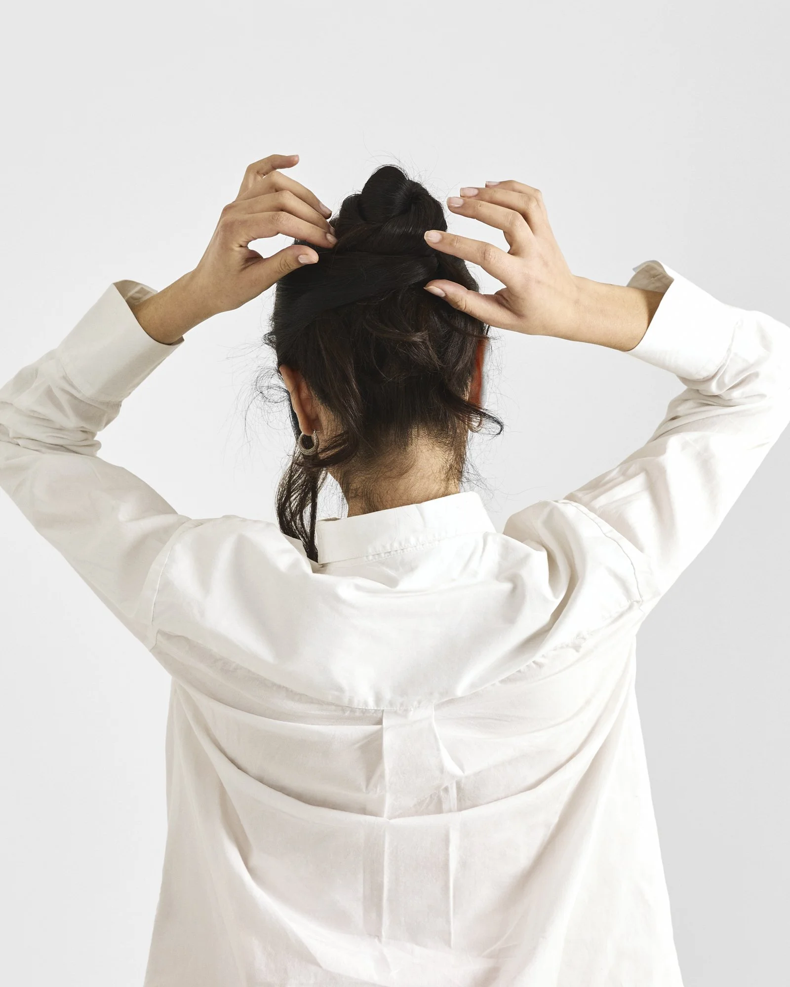 A person with dark hair styled in a loose bun, wearing a white long-sleeve shirt, is adjusting their hair with both hands against a plain white background.