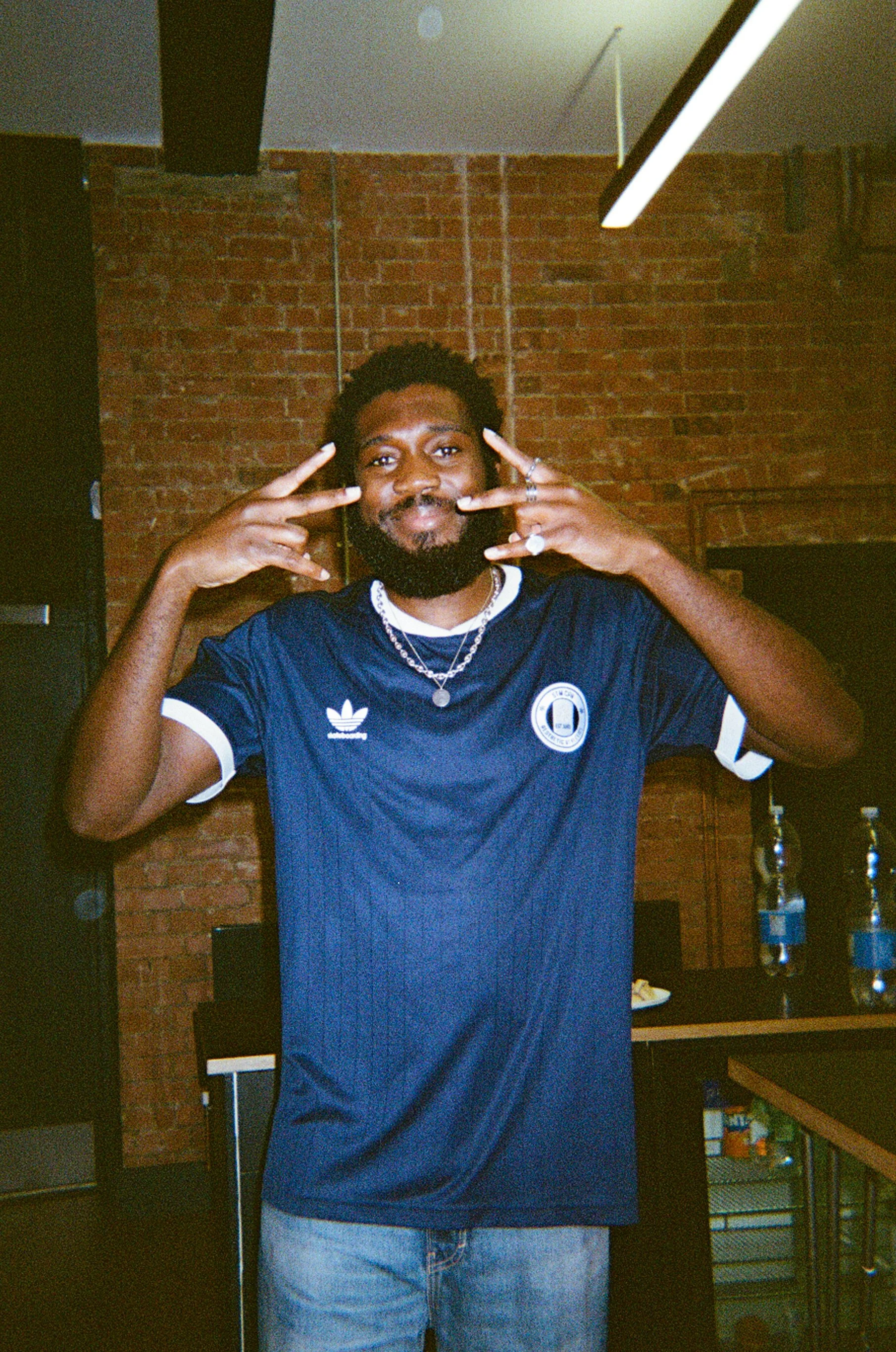 A young man with a beard and curly hair smiling and making peace signs with both hands. He is wearing a navy blue sports jersey with white accents and light blue jeans, standing indoors with a brick wall in the background.